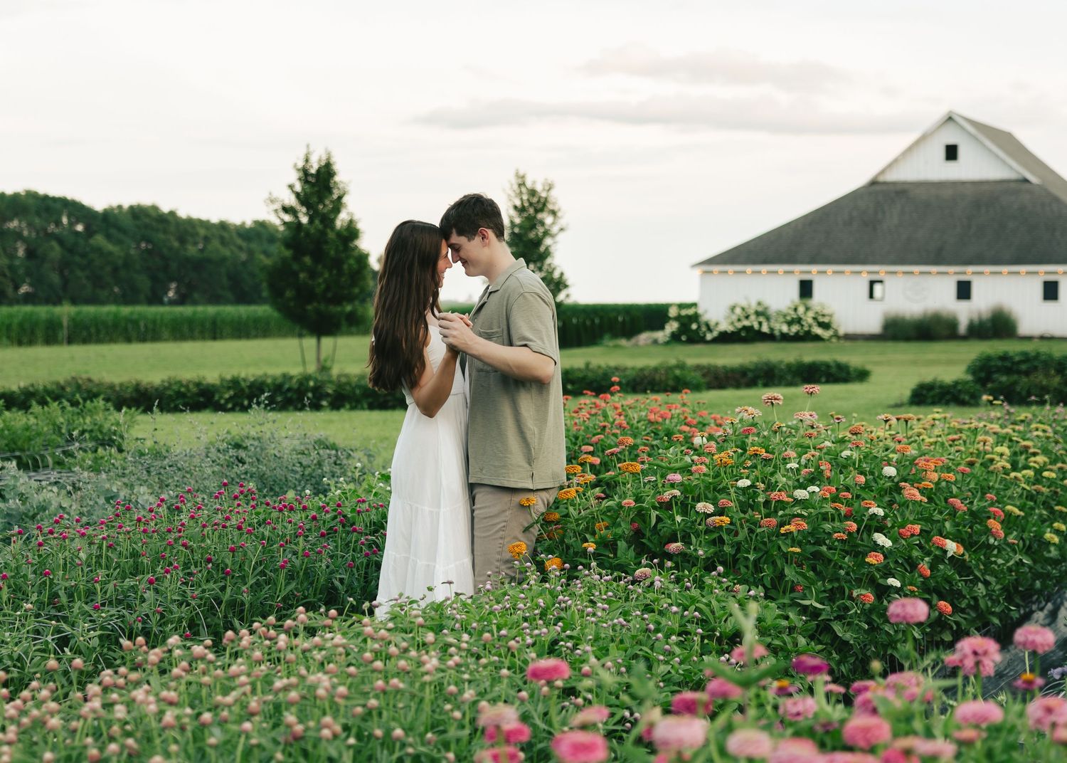 Couple embracing in a vibrant flower garden with a white barn in the background on a lush green farm.