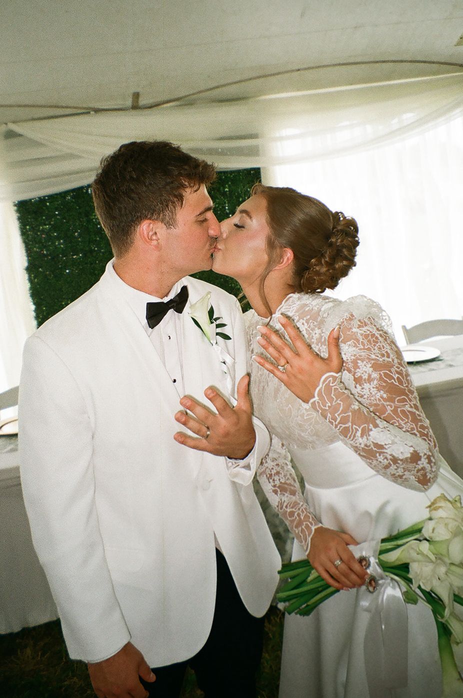 Bride and groom sharing a kiss at their wedding reception, wearing white lace gown and white tuxedo with black bow tie.