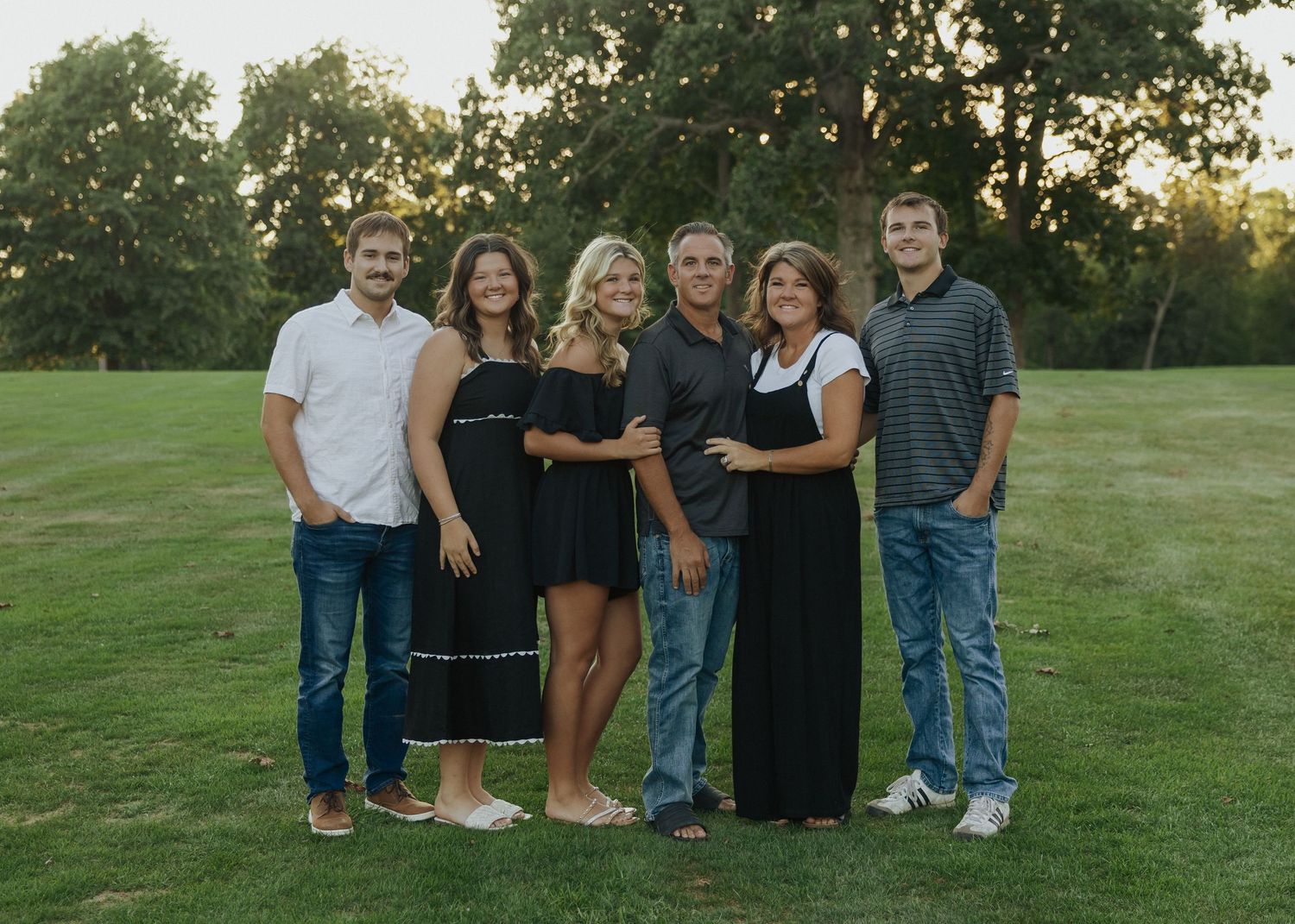 Family of six posing together on a green lawn with trees in the background during a golden hour outdoor photo session.