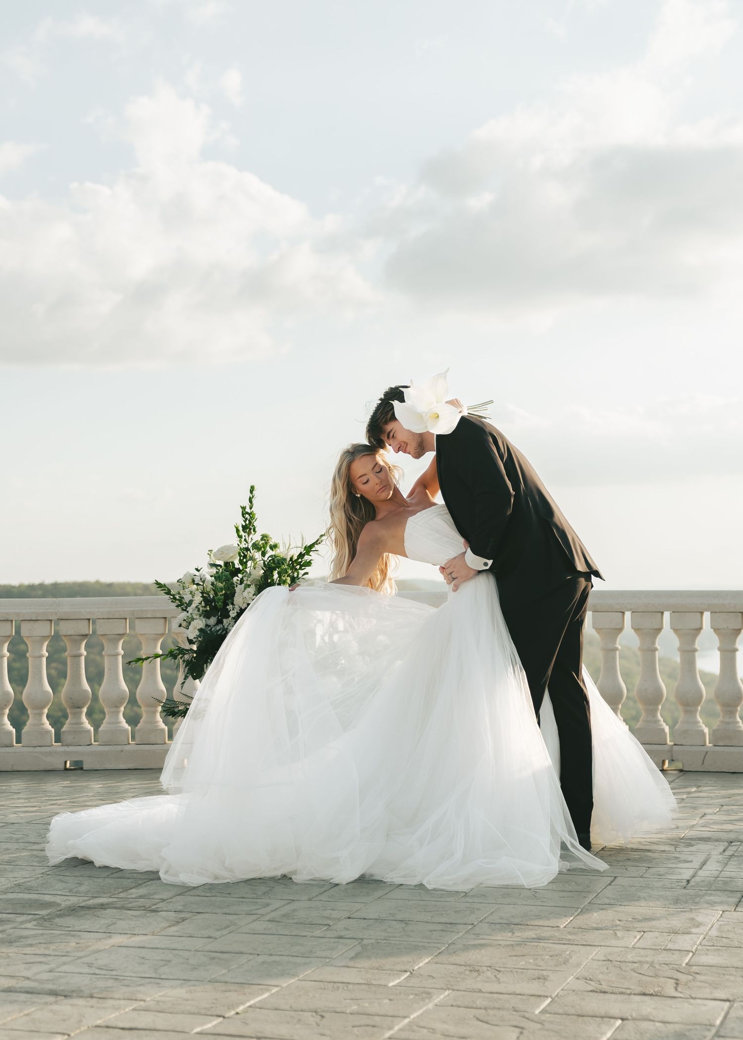 Bride in white ballgown and groom in black tuxedo share romantic kiss on elegant stone terrace under cloudy sky.