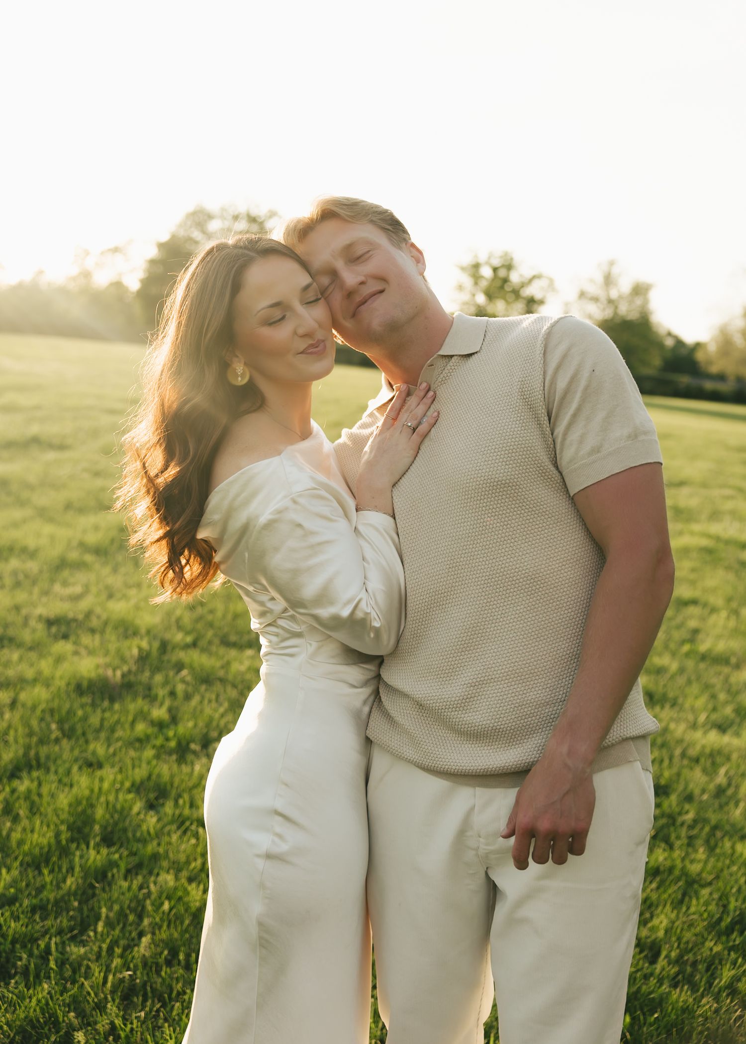A woman leans against a smiling man in a warmly lit green field during golden hour.