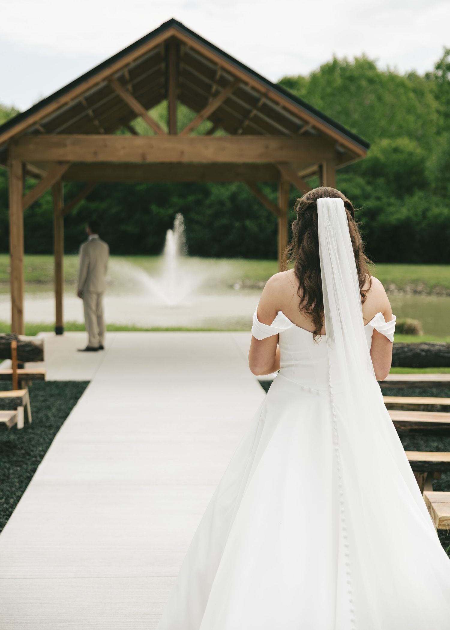 Bride in off-shoulder gown with veil walks toward groom near a wooden pavilion with fountain backdrop.