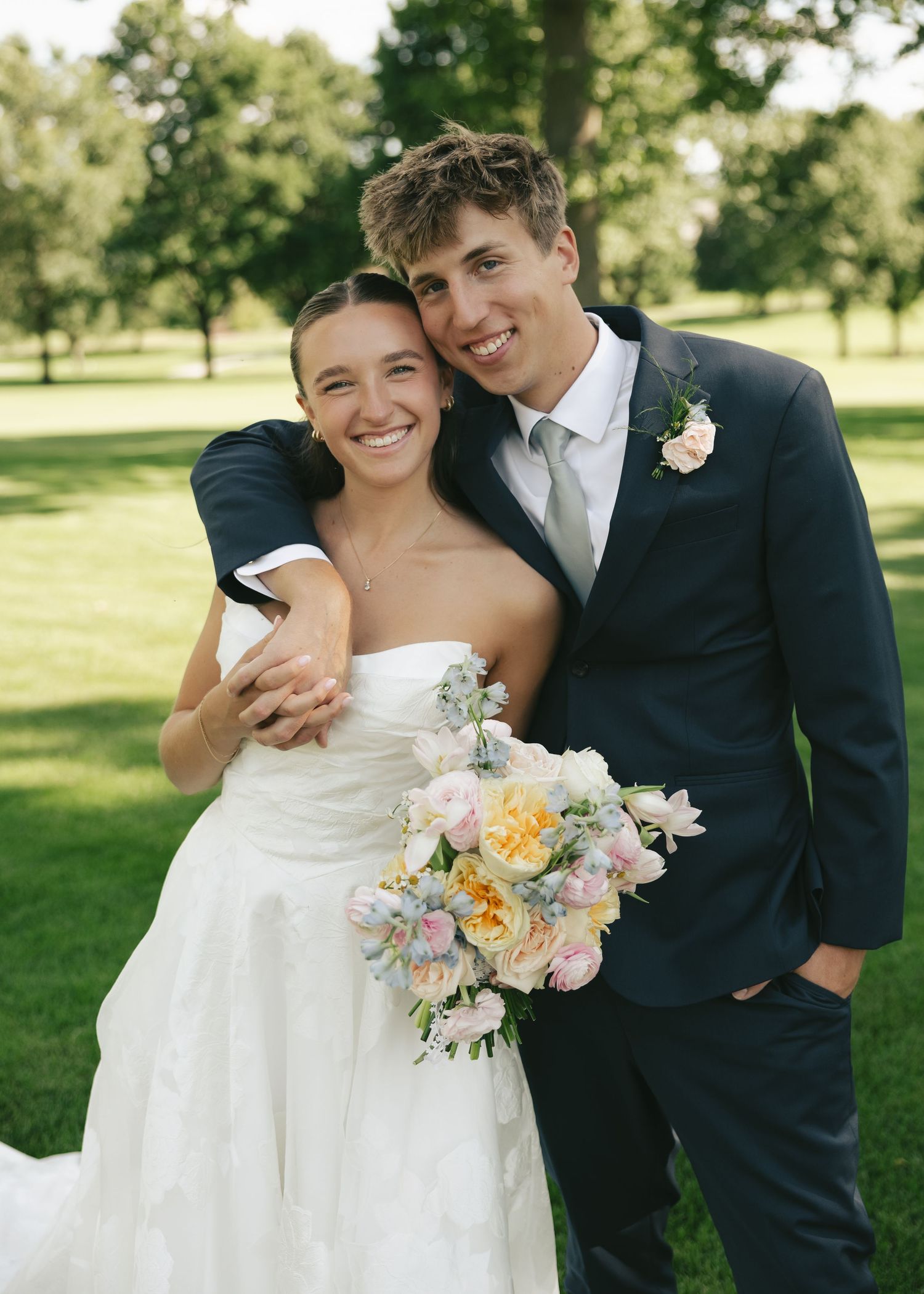 Smiling bride and groom pose outdoors on wedding day, she holds pastel bouquet as he embraces her shoulders.