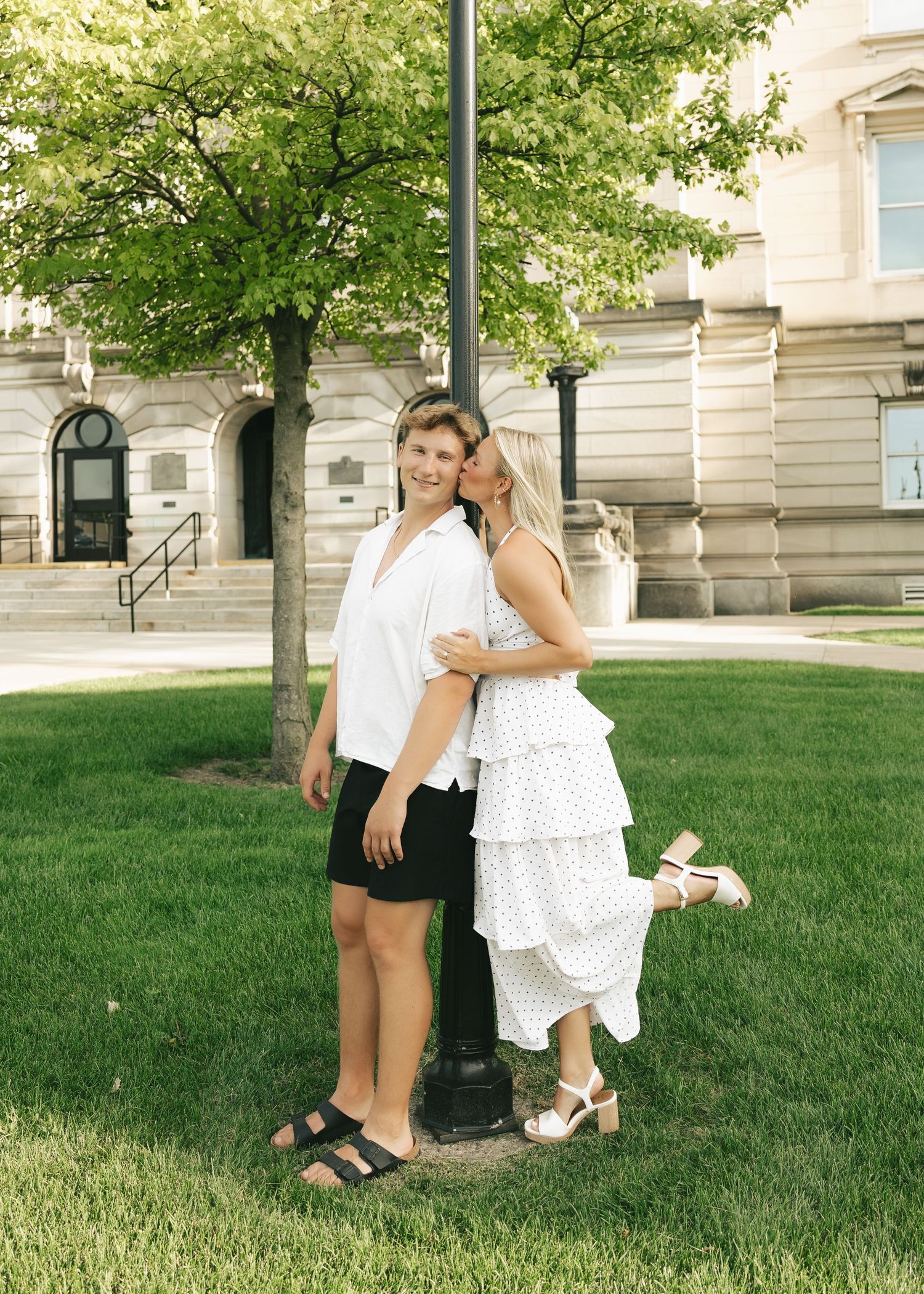 A couple poses together on green grass near a lamppost outside a historic building on a sunny day.