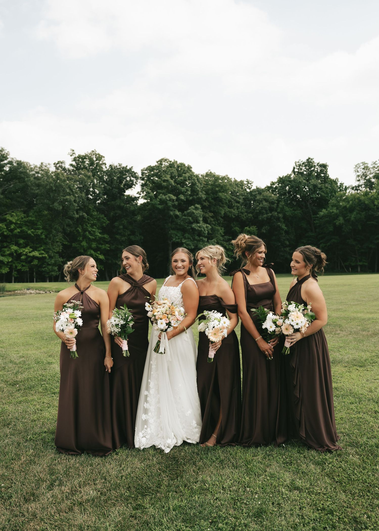 Bride in white gown stands with five bridesmaids in brown dresses holding white bouquets on a green lawn.