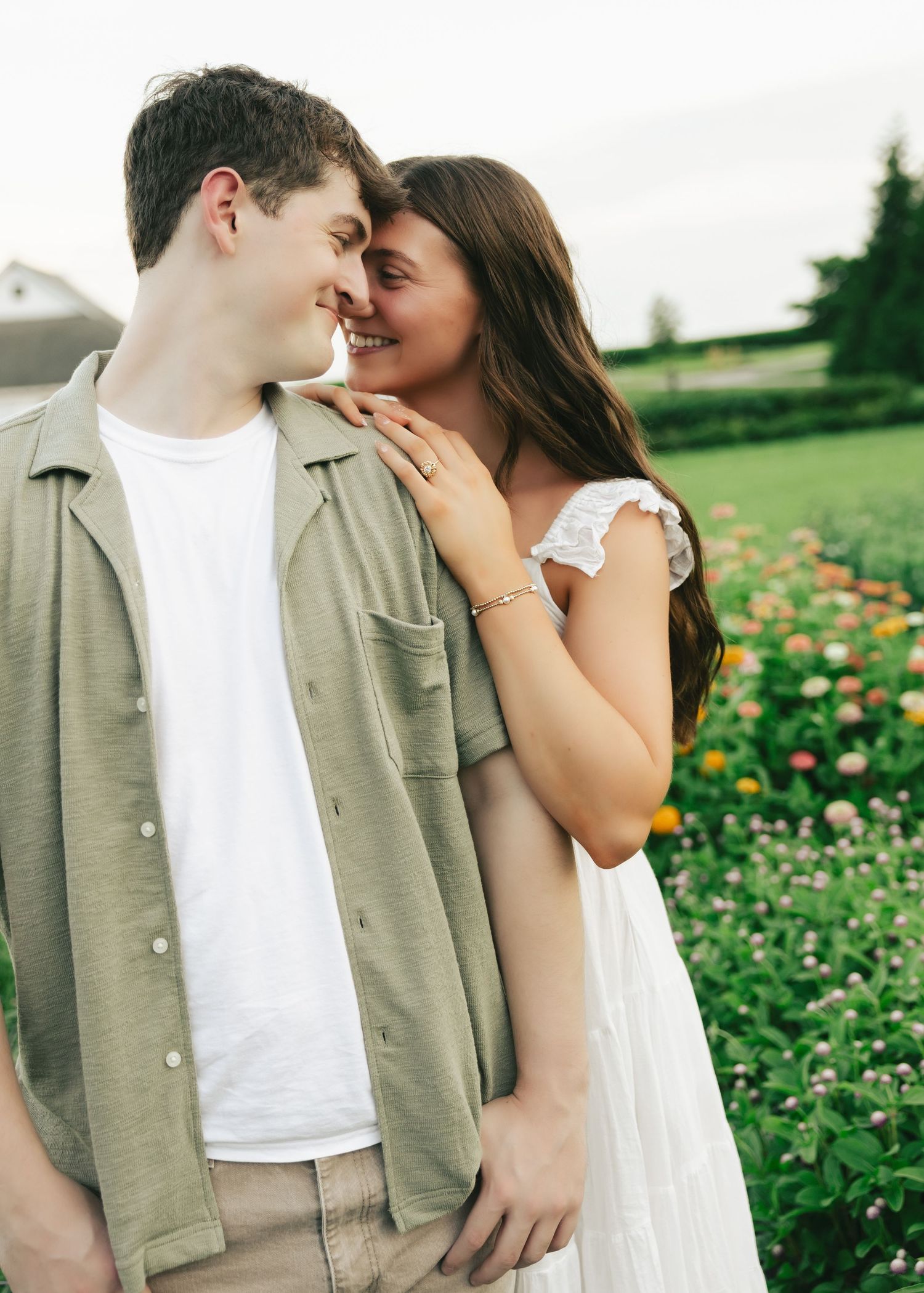 A young couple smiles and embraces near a colorful flower garden, the woman in white dress and man in sage green shirt.