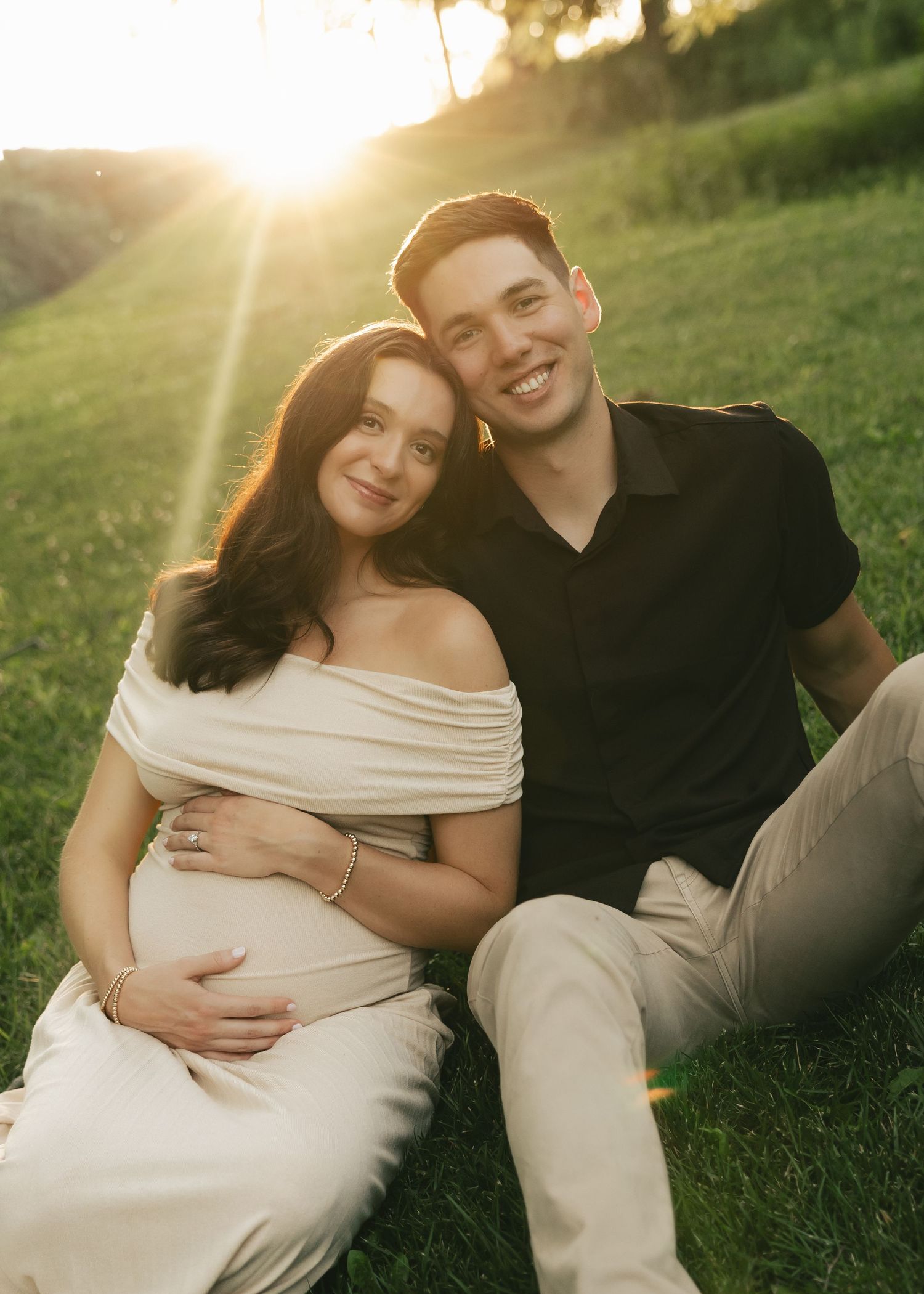 Couple sitting on grass at golden hour, pregnant woman in white off-shoulder dress, man in black shirt smiling warmly.