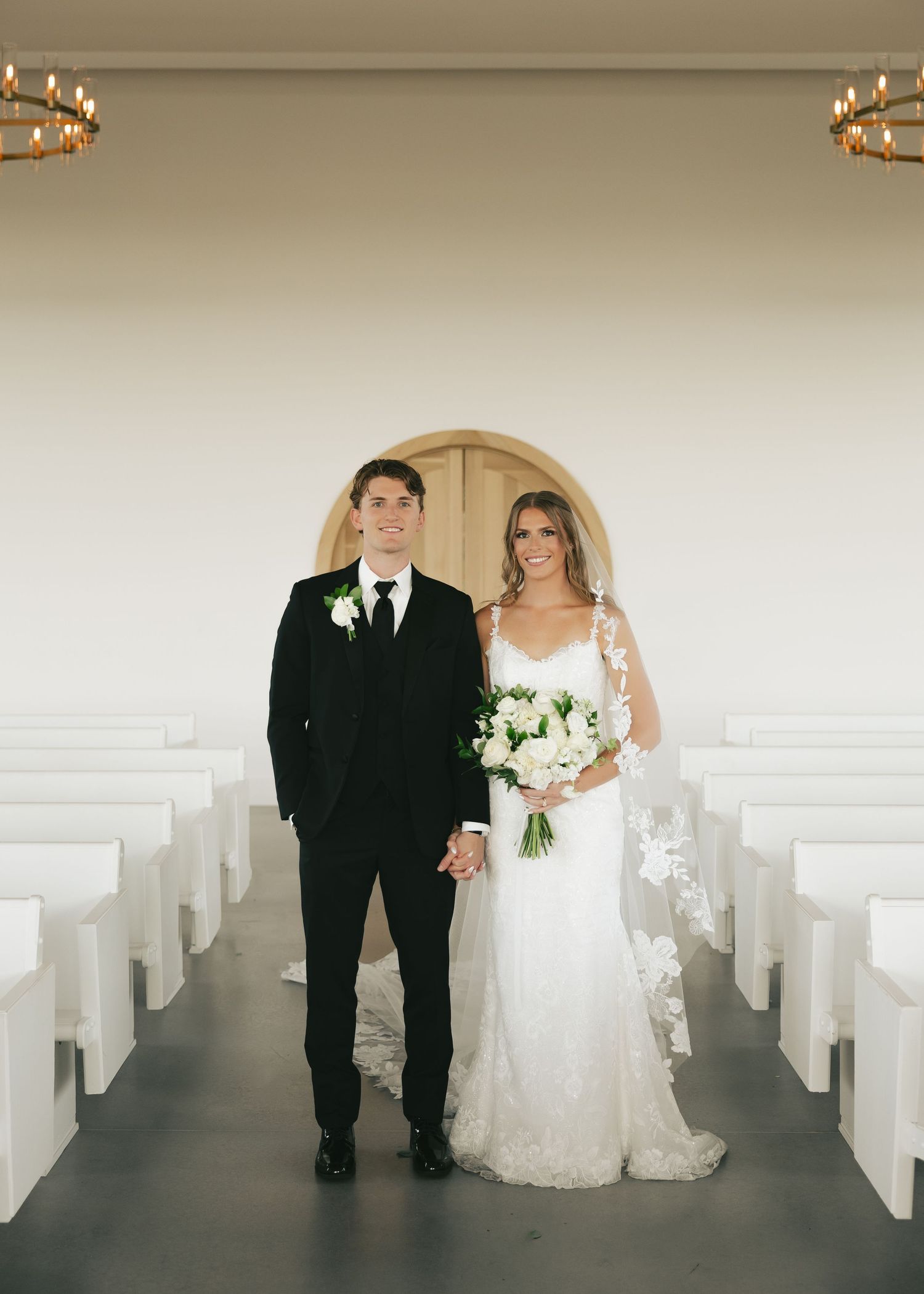 Bride and groom holding hands in a modern white chapel aisle, bride in lace gown with bouquet, groom in black tuxedo.