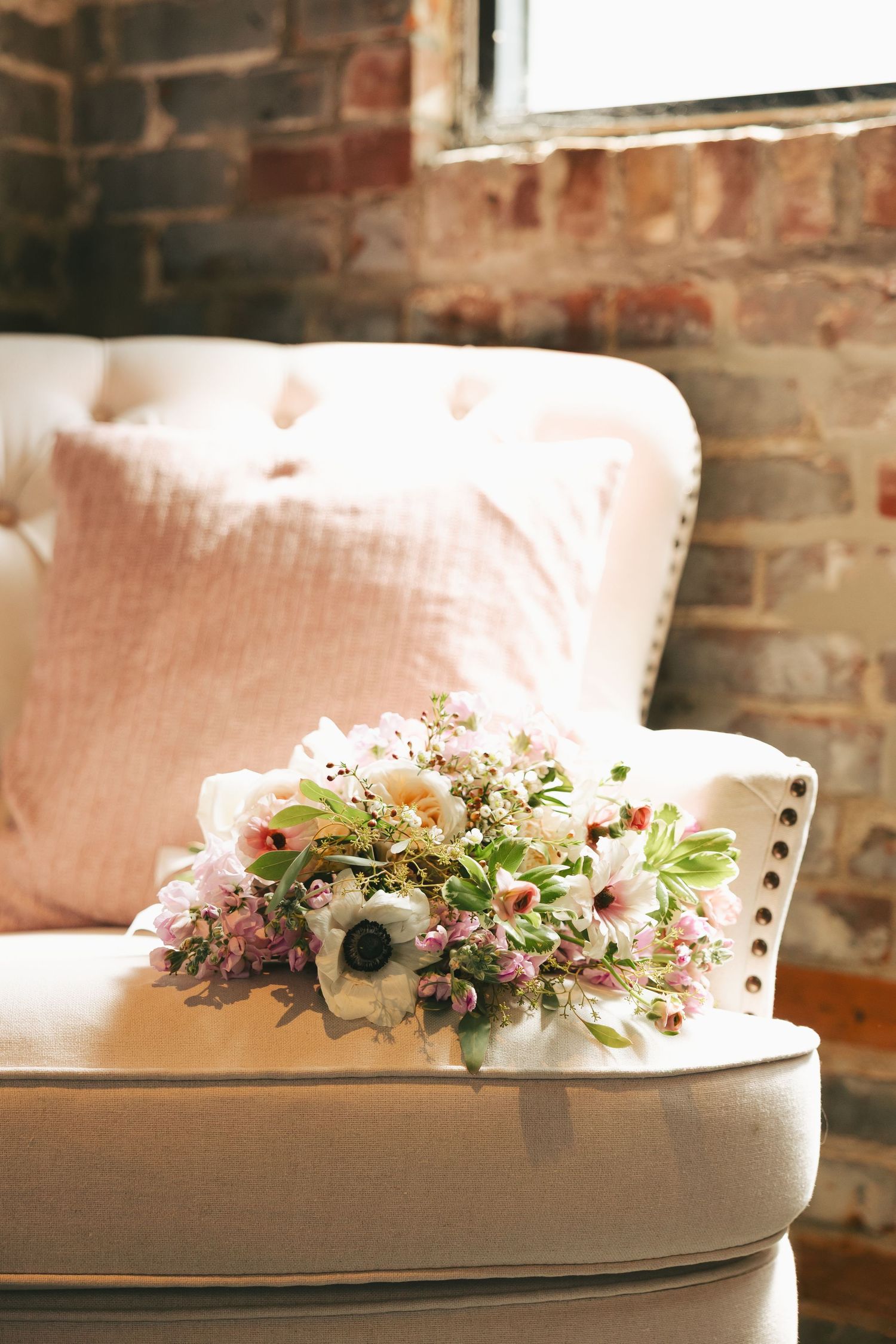Elegant bridal bouquet of anemones and wildflowers resting on tufted cream sofa near brick wall.