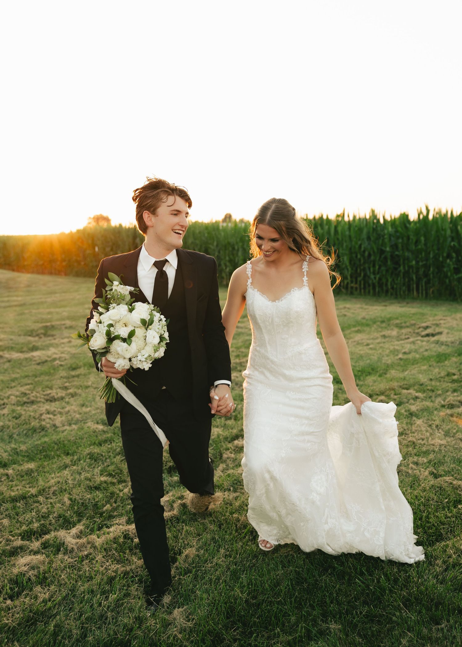 Bride and groom walk hand-in-hand through a green field at golden hour, bride in lace gown holding her dress.