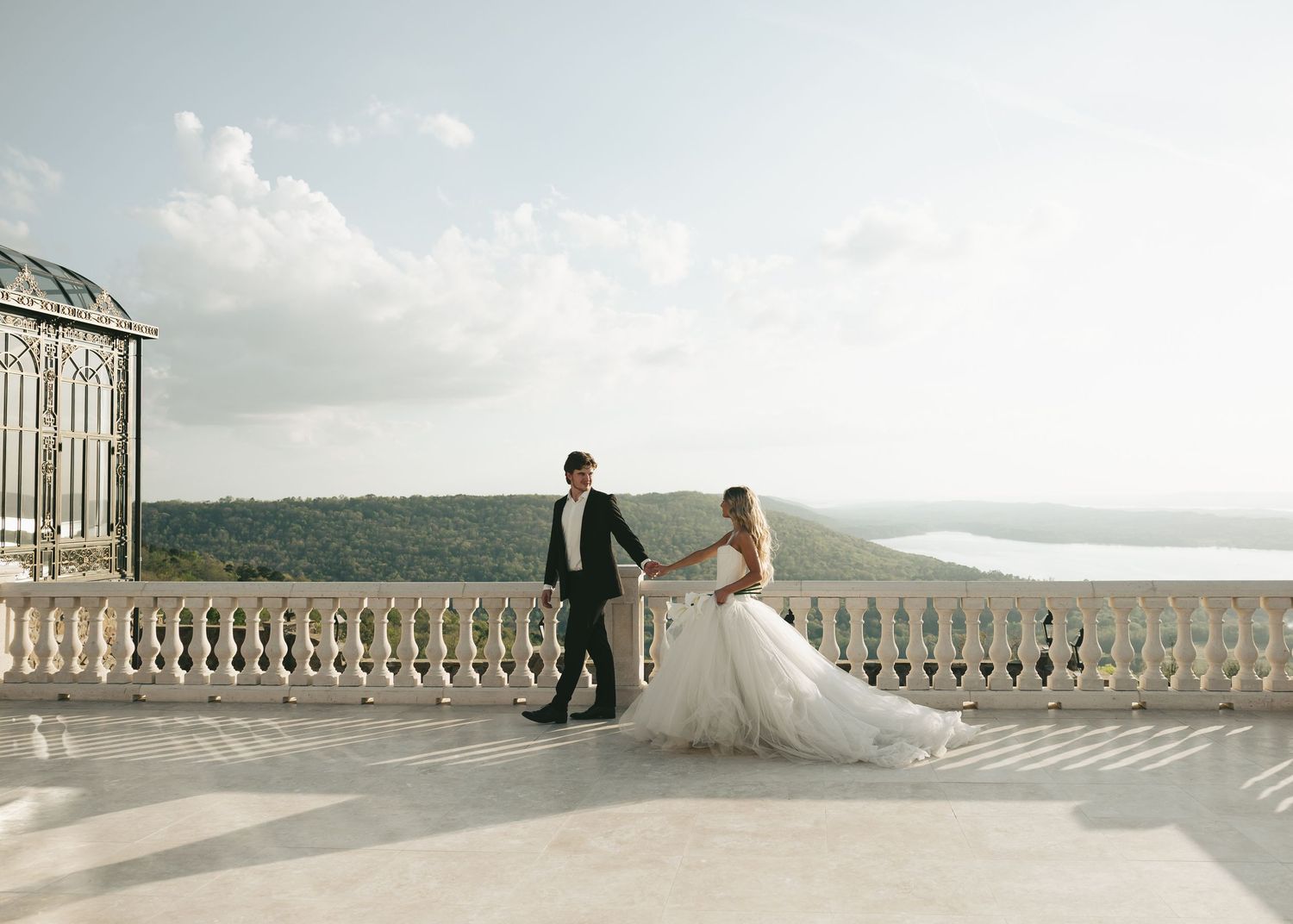 Bride and groom hold hands on a grand terrace with sweeping scenic views on their wedding day.