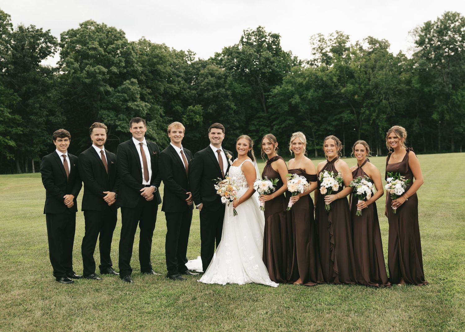 Wedding party posing outdoors, groomsmen in black suits and bridesmaids in brown gowns with bride and groom centered.