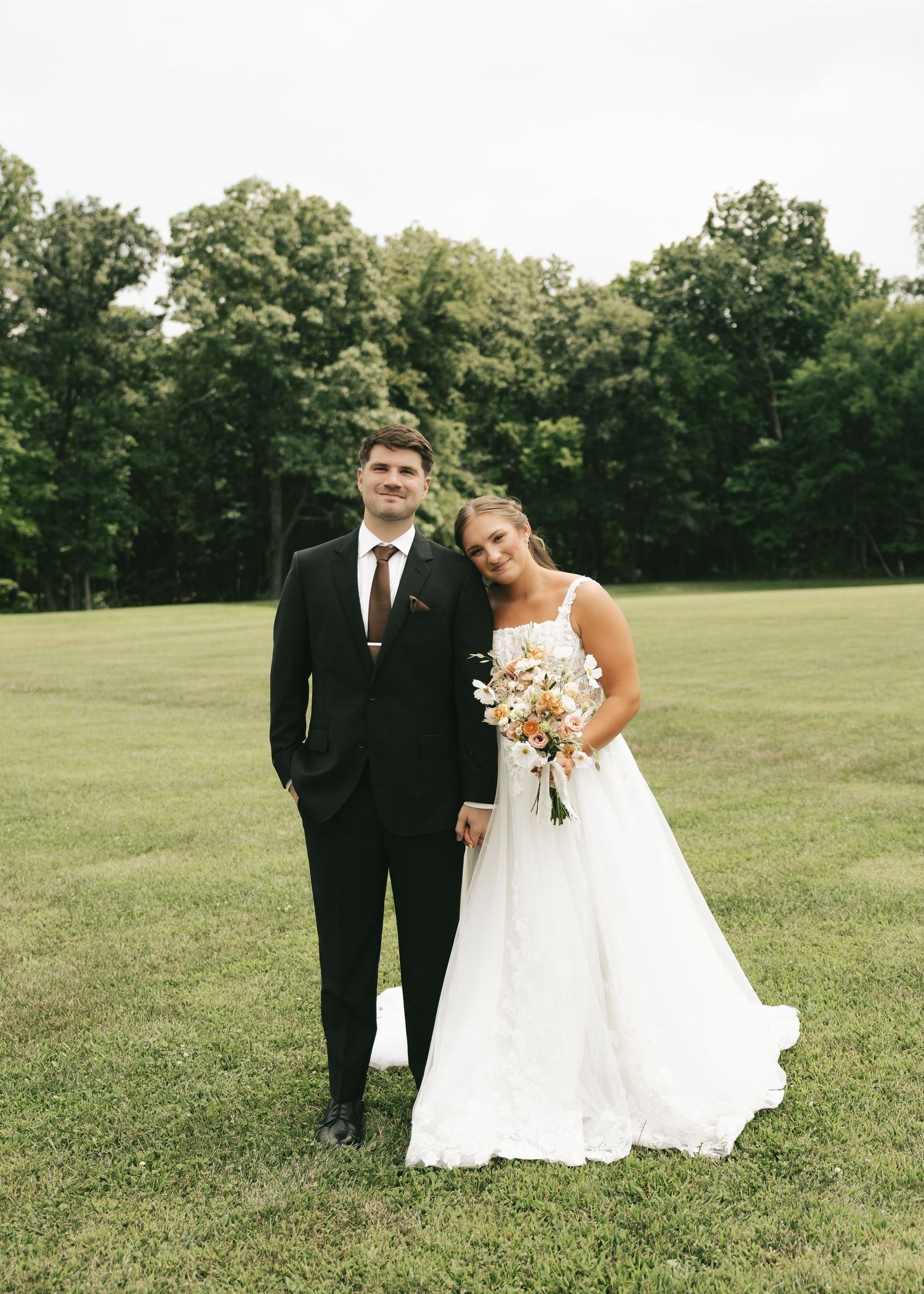 Bride in white gown holding floral bouquet stands with groom in black suit on green lawn with trees.