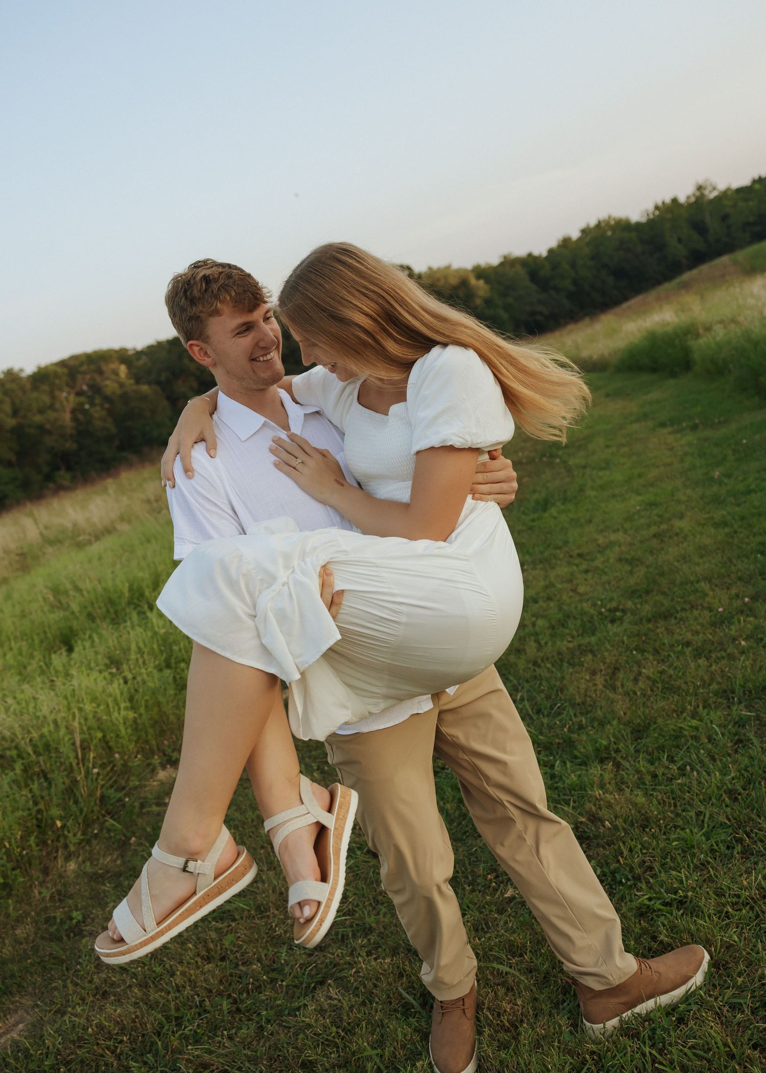 Couple in white outfits laughing and spinning together in a sunlit green meadow.