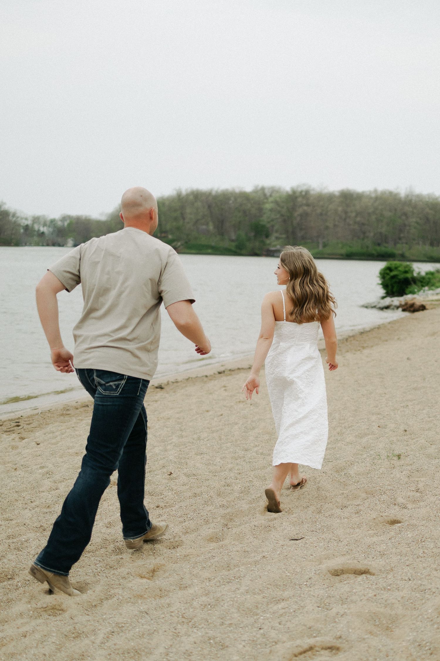 Couple walking hand in hand along a sandy lakeside beach, woman in white sundress during engagement photos.
