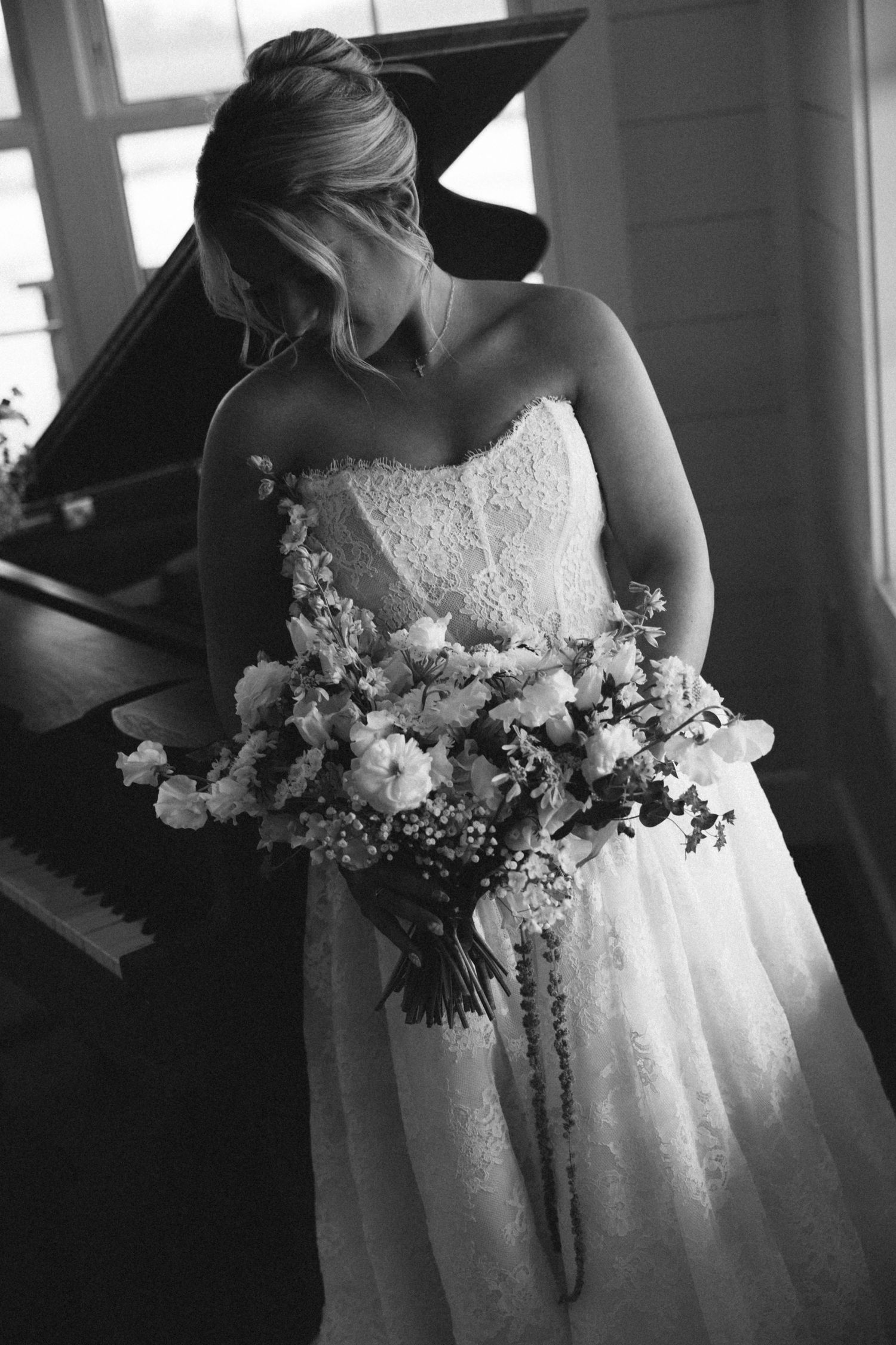 Black and white photo of a bride in strapless lace gown holding a cascading bouquet near a grand piano.