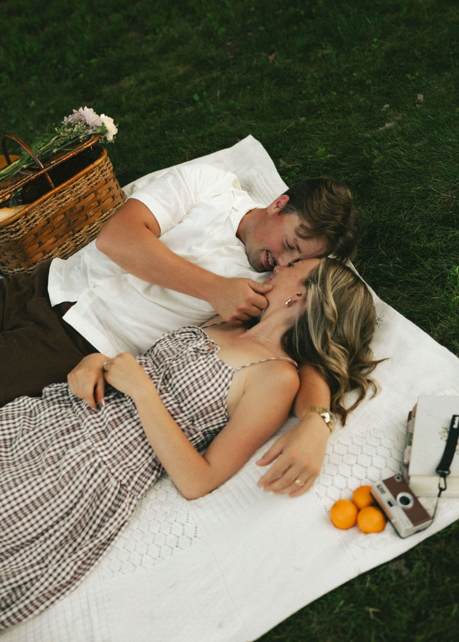 Couple sharing a romantic picnic on a white blanket in green grass with a wicker basket and oranges nearby.