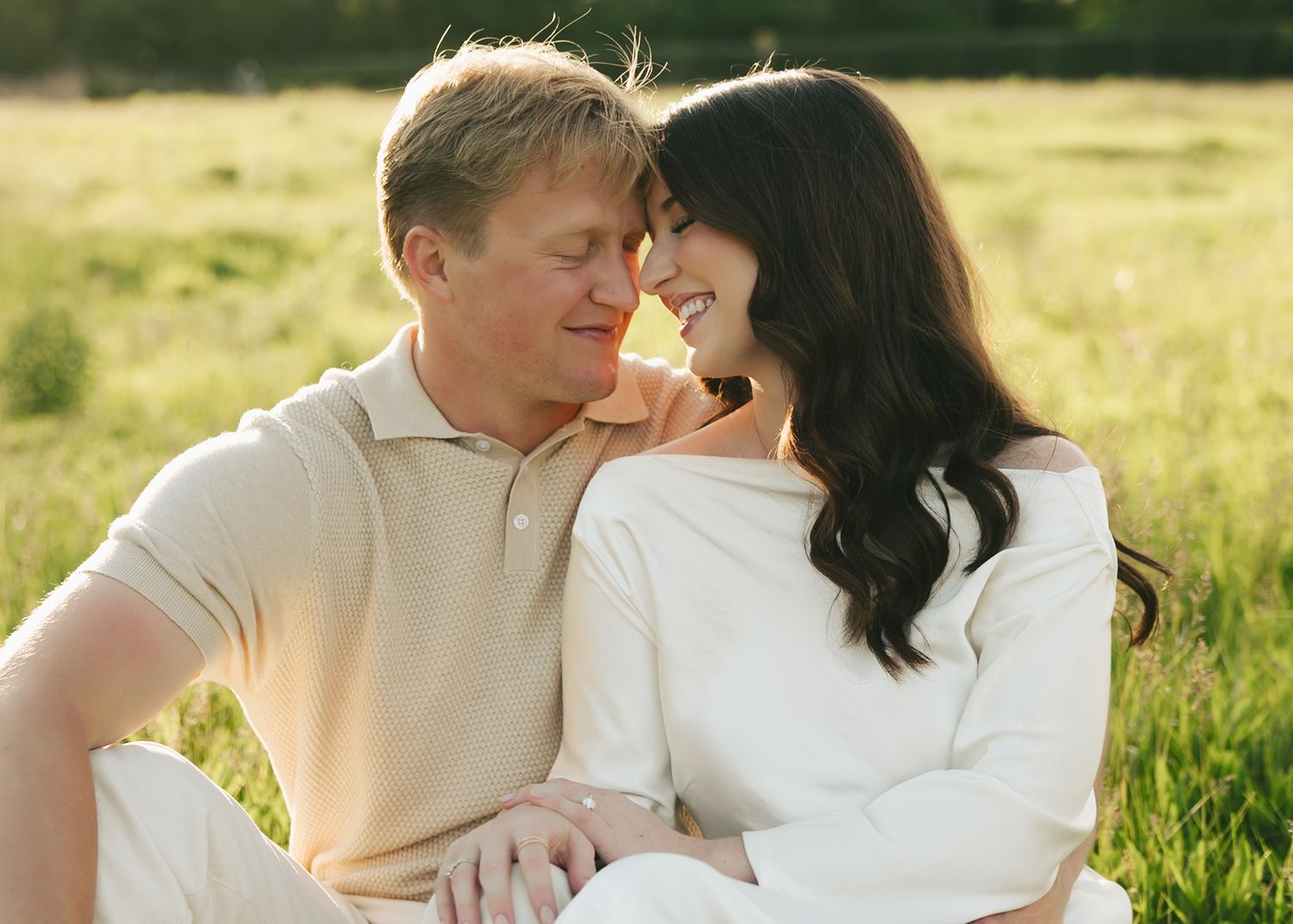 Couple sitting in a sunlit green field, sharing a tender romantic moment during an outdoor engagement session.