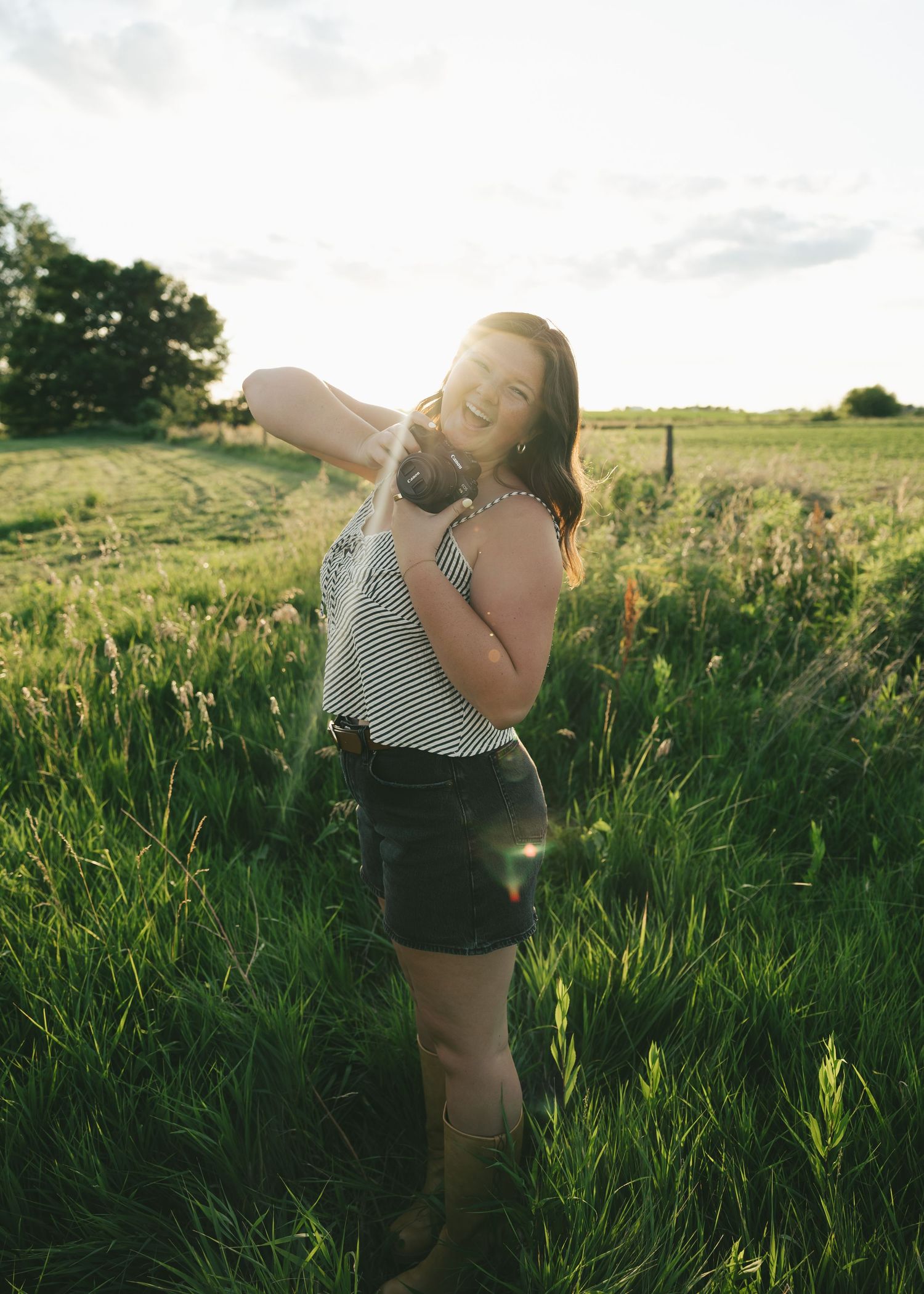 Young woman in striped top and denim shorts standing in lush green field at golden hour sunset.