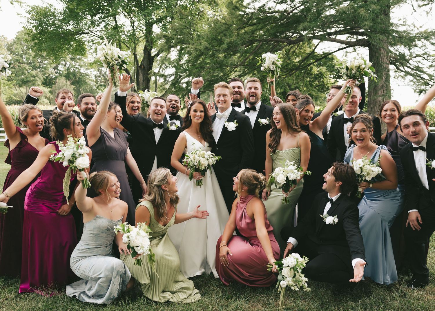 A joyful wedding party celebrates outdoors, with bride and groom surrounded by bridesmaids and groomsmen holding white bouquets.