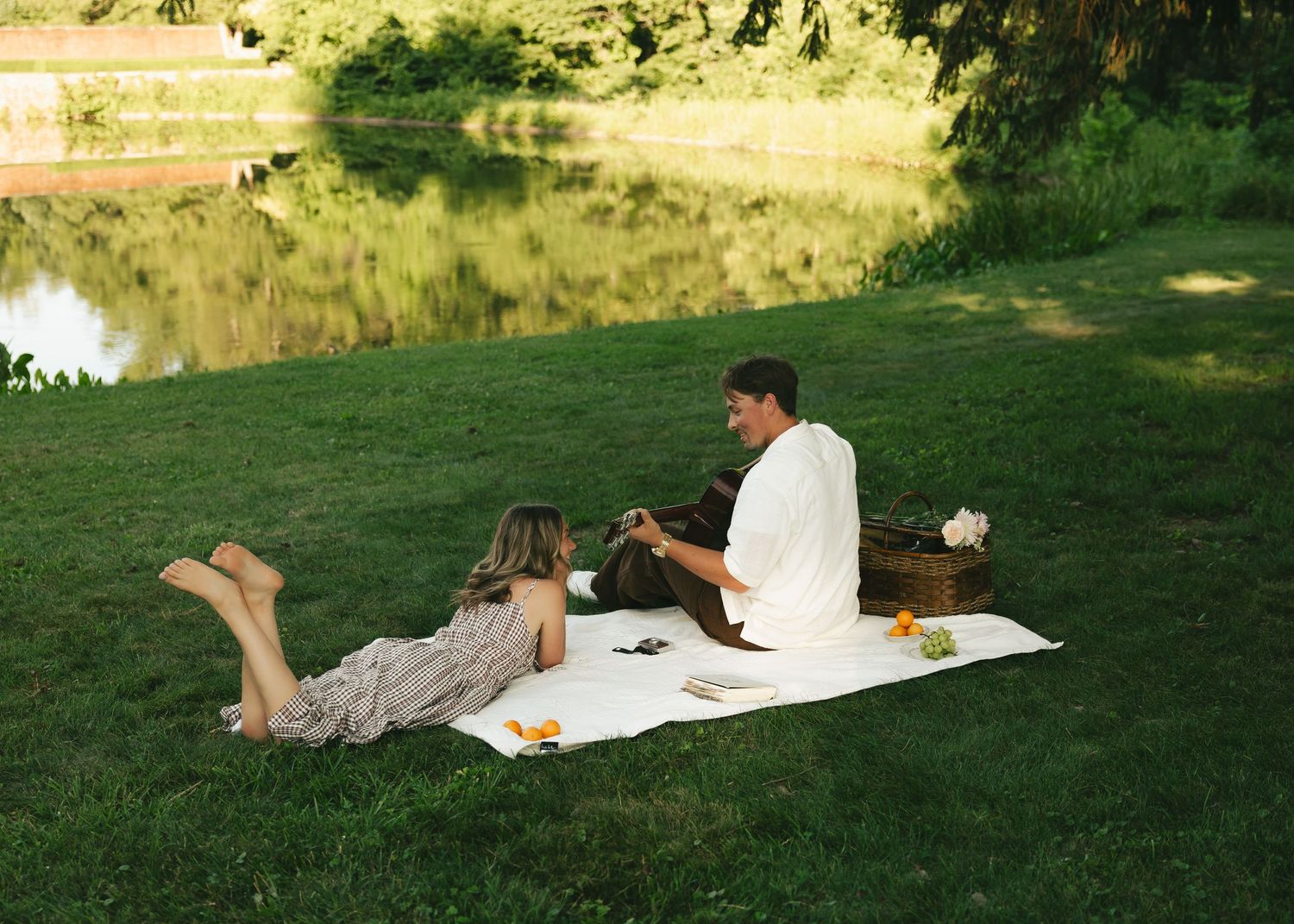 Couple enjoying a romantic picnic on a green lawn by a tranquil river, with a wicker basket nearby.