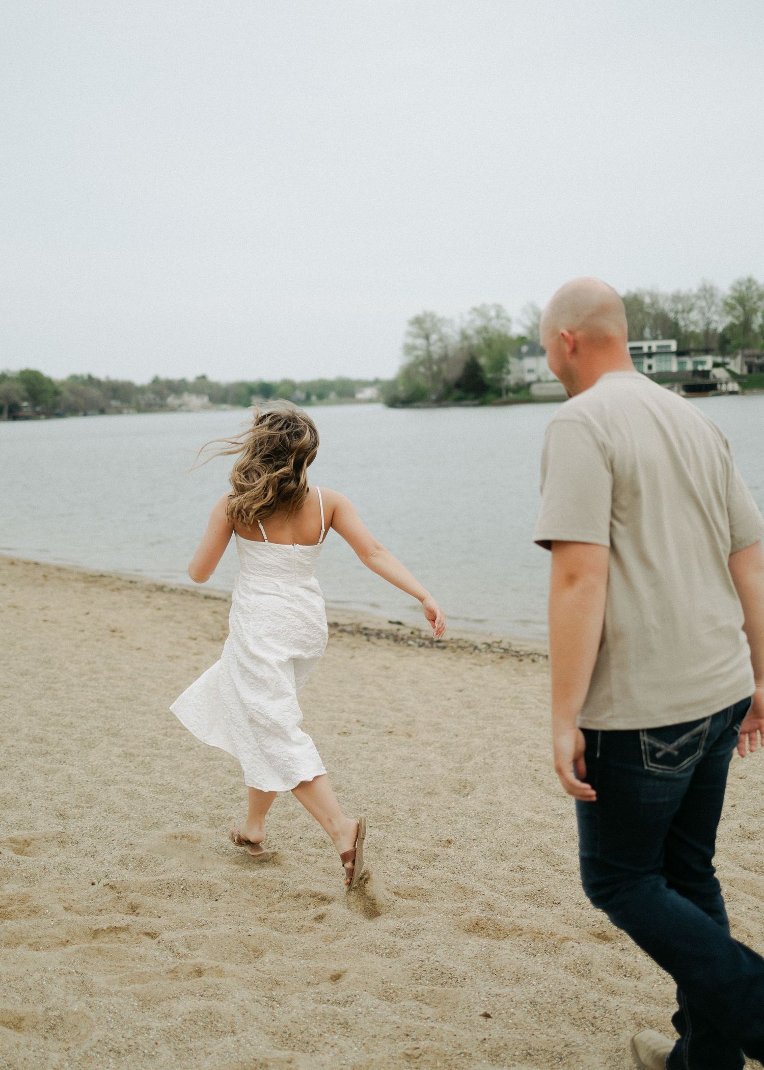 Couple walking barefoot on a sandy beach near a calm lake on an overcast day.