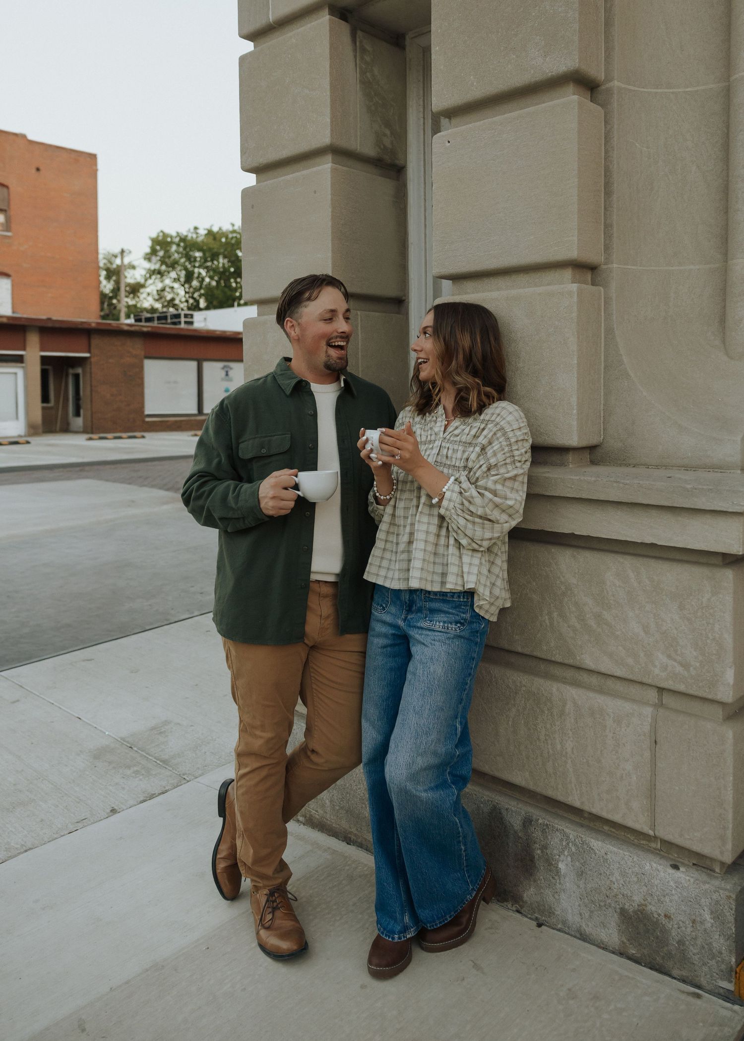Happy couple laughing and holding coffee cups outside a stone building on a casual date.