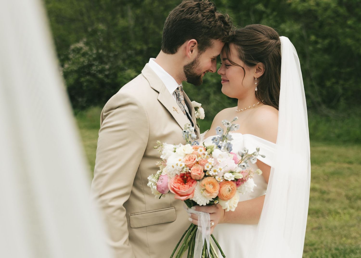 Newlyweds share an intimate forehead touch moment outdoors, bride holding a vibrant spring wedding bouquet in white dress.