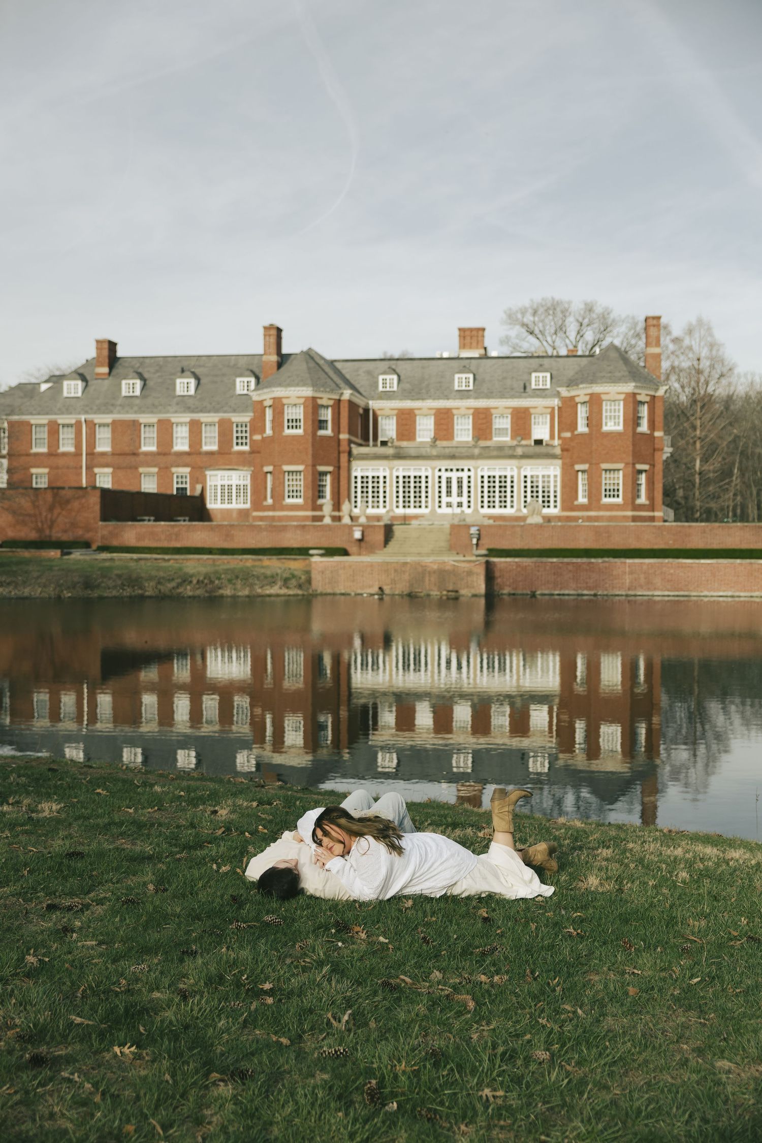 Couple lying on grass by reflective pond with grand brick estate mansion in background.