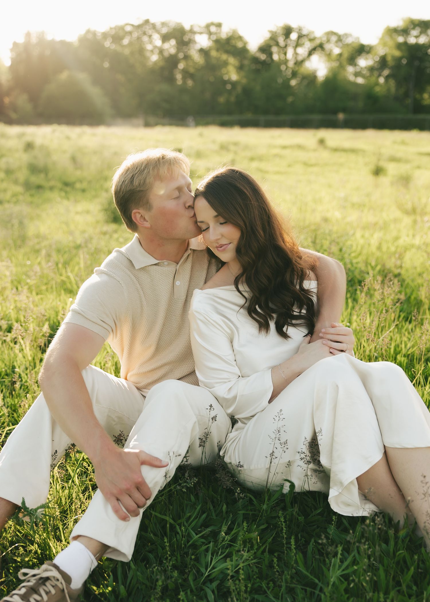 Couple sits together in sunlit meadow, man kissing womans cheek as golden light filters through the trees.