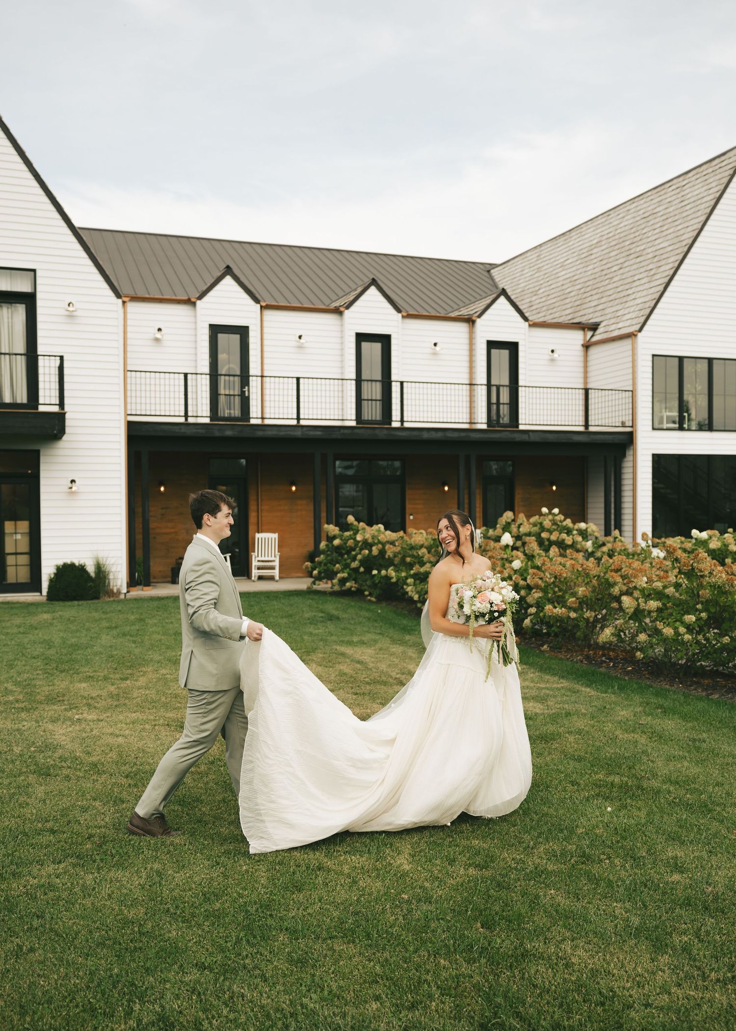 Bride and groom walk across green lawn outside white farmhouse wedding venue, groom carrying bride's flowing white train.