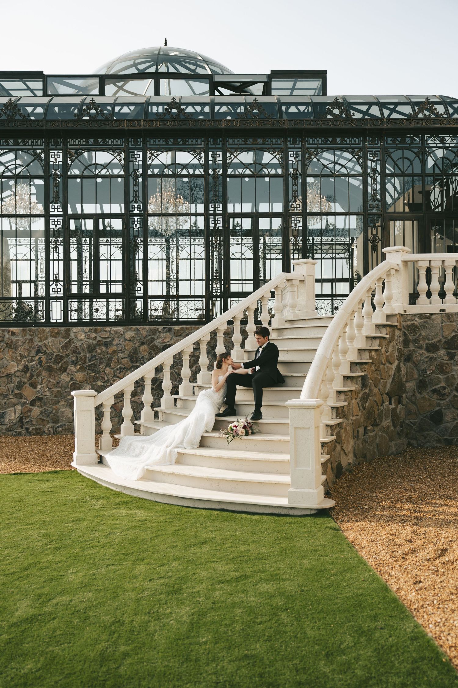 Bride and groom seated on ornate white staircase with dramatic iron greenhouse structure backdrop.