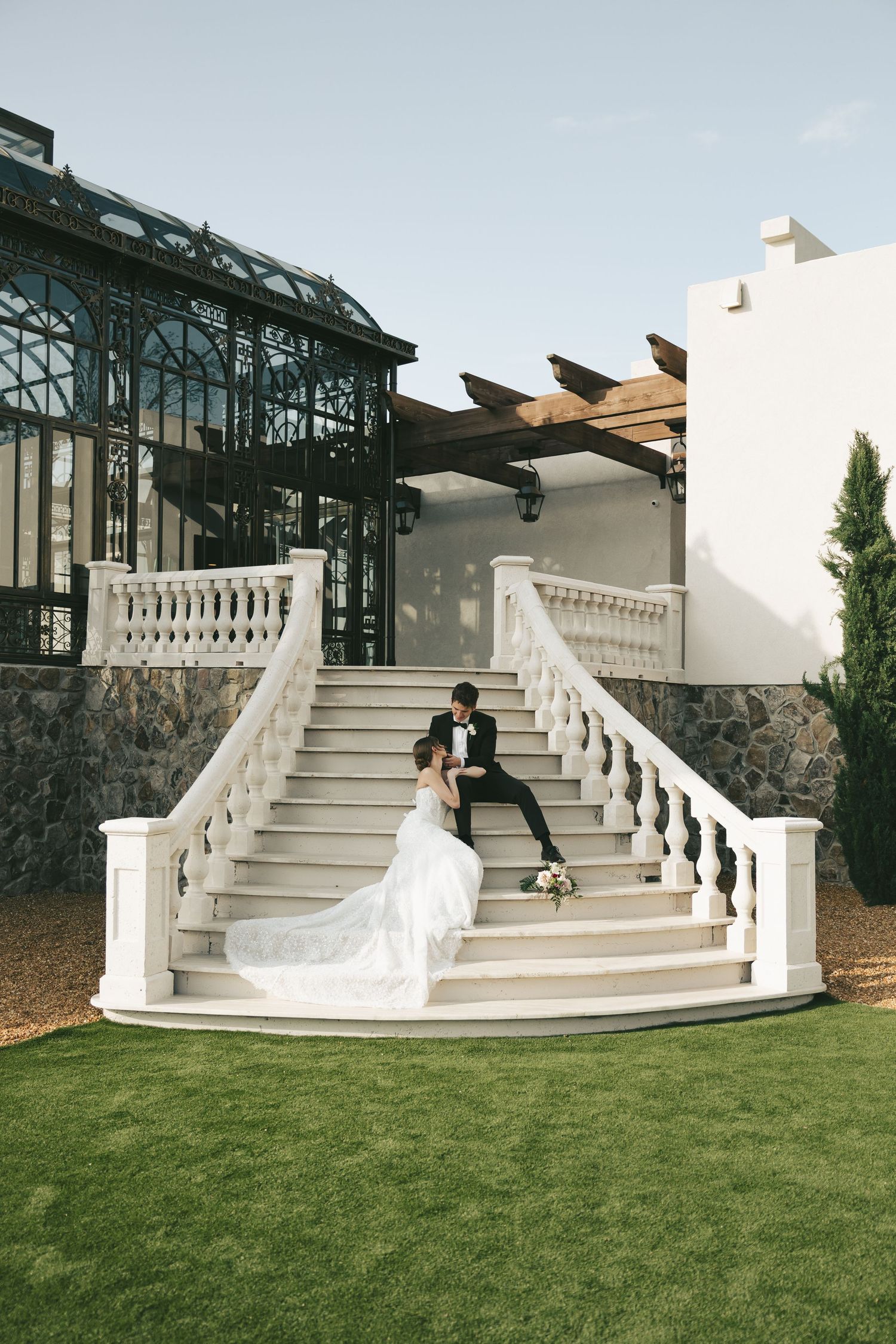 Bride and groom sitting on grand white staircase outside elegant greenhouse venue on wedding day.