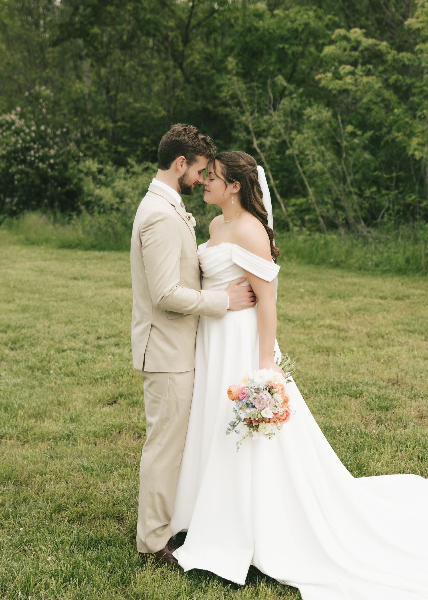 Bride and groom in beige suit share a romantic moment in a lush green meadow on their wedding day.