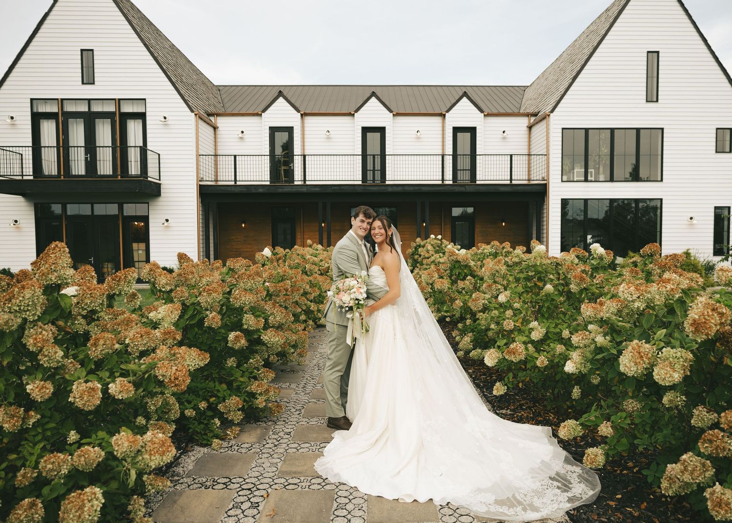Bride and groom pose among lush hydrangea gardens in front of a white modern farmhouse wedding venue.