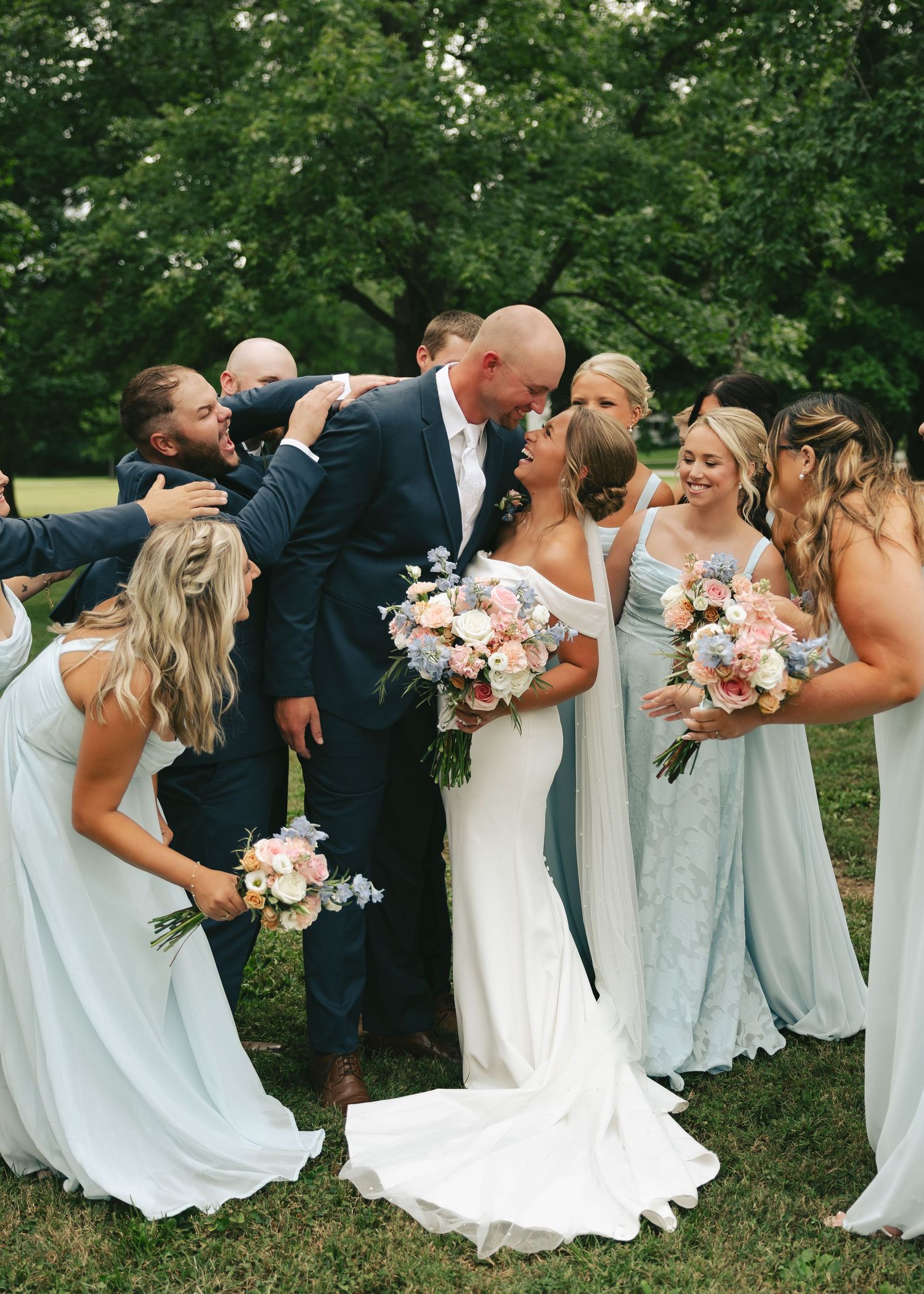 Wedding party surrounds bride and groom sharing a kiss outdoors, bridesmaids in blue gowns holding floral bouquets.