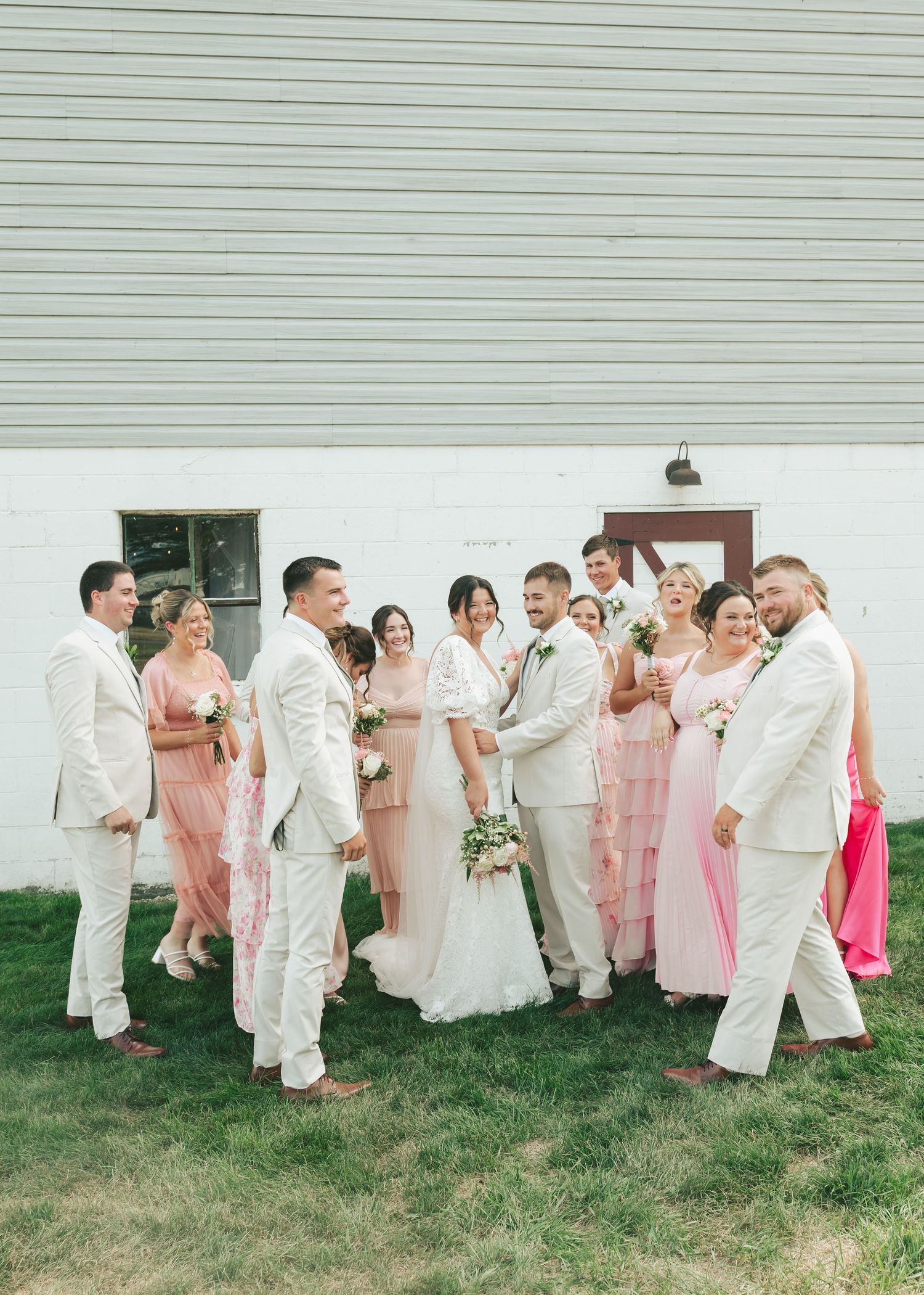 Wedding party in pink and white attire laughing together outside white barn venue.