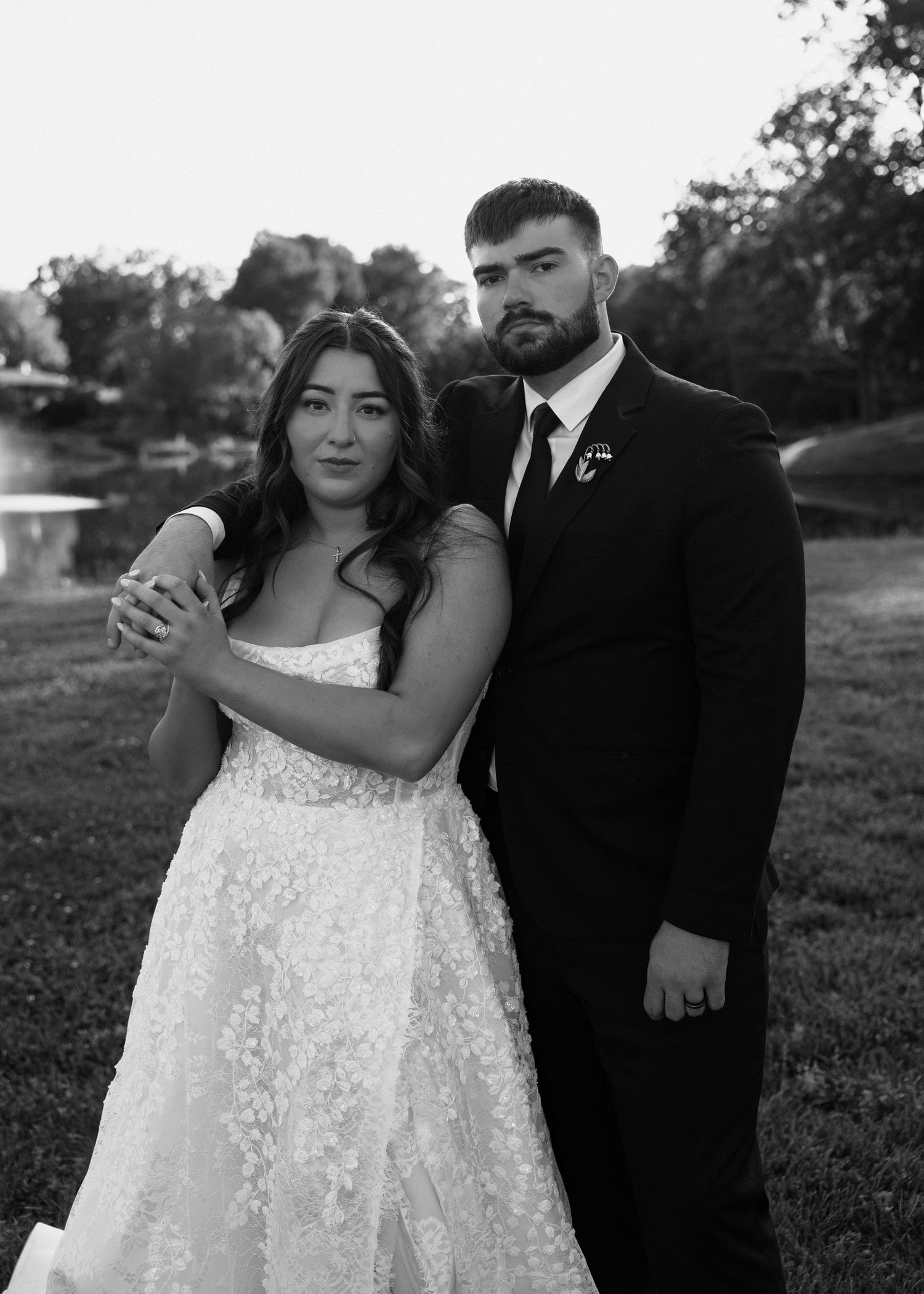 Black and white portrait of bride and groom embracing outdoors near a pond, groom in black suit, bride in lace gown.