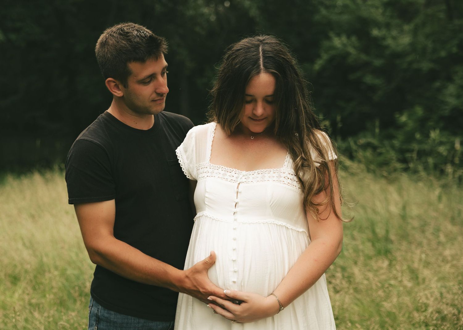 A couple tenderly cradles a pregnant belly outdoors in a lush green field during a maternity photo session.