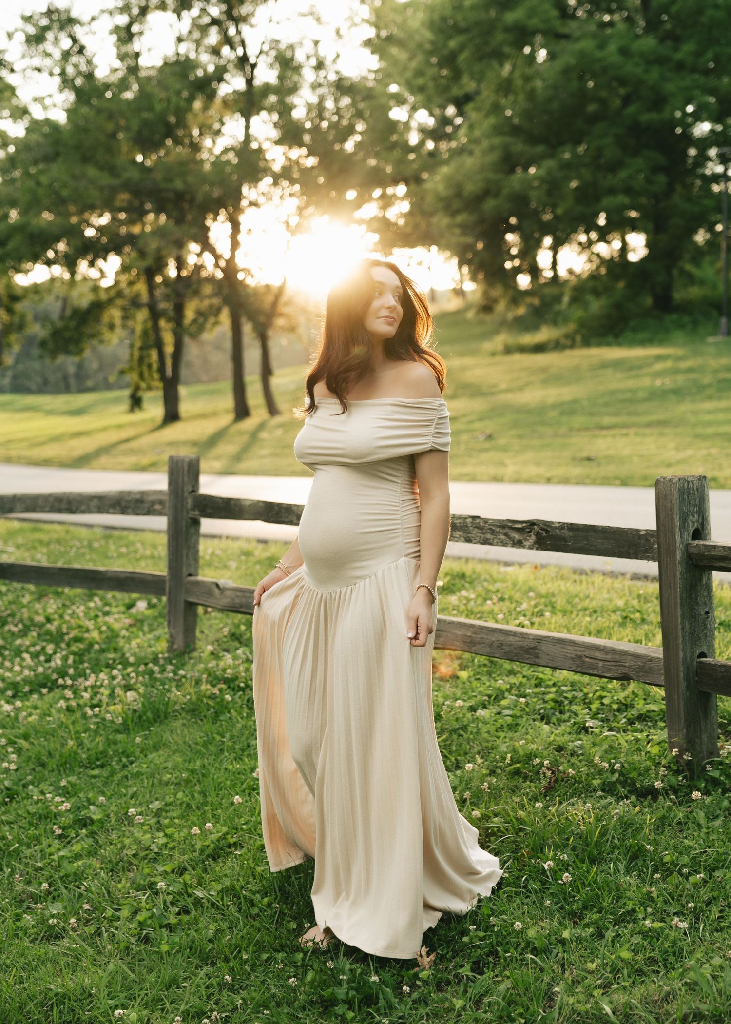 Pregnant woman in elegant off-shoulder cream gown standing by rustic fence at golden hour sunset.