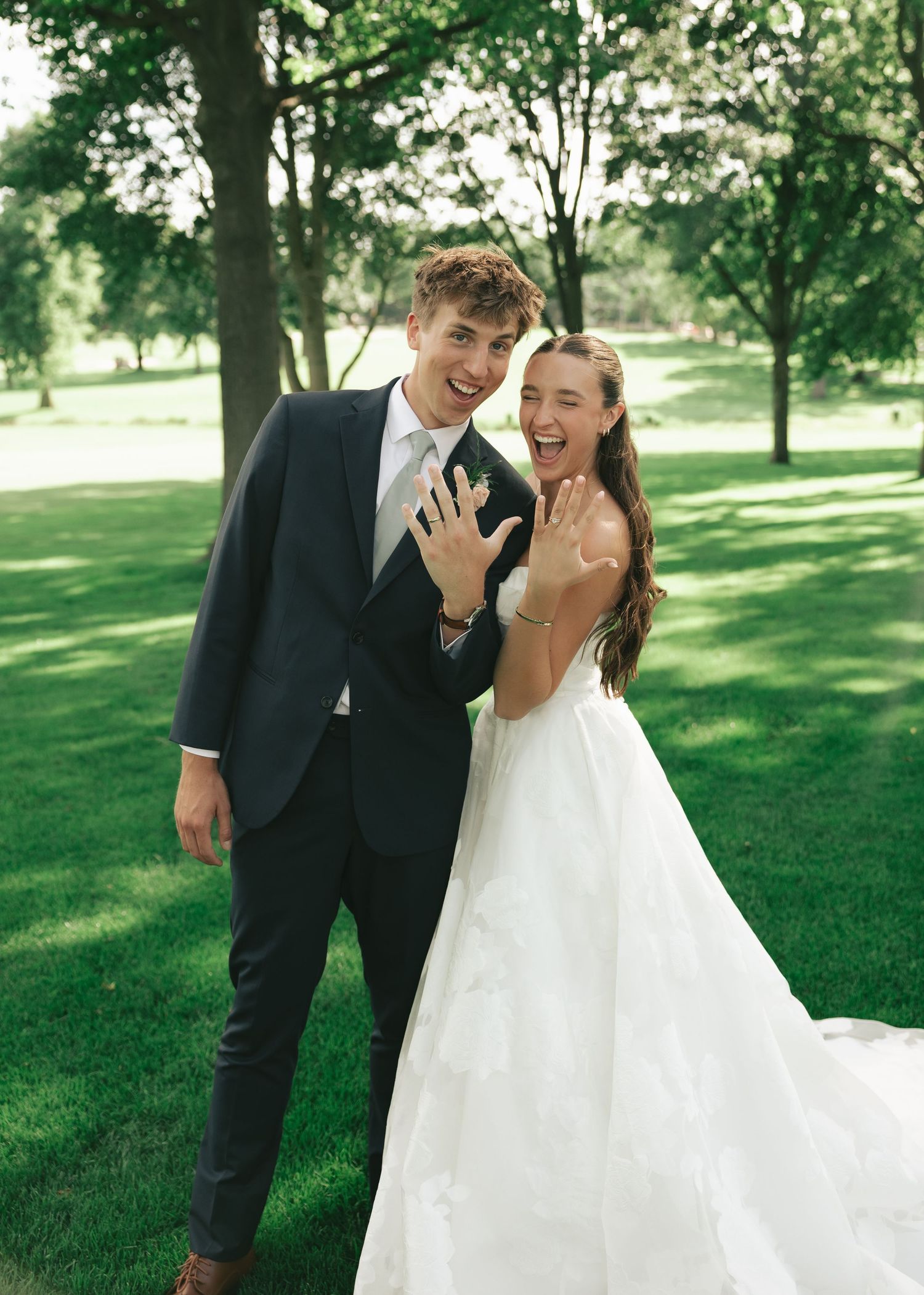 A joyful newlywed couple in a park, showing off their wedding rings, groom in navy suit, bride in white gown.