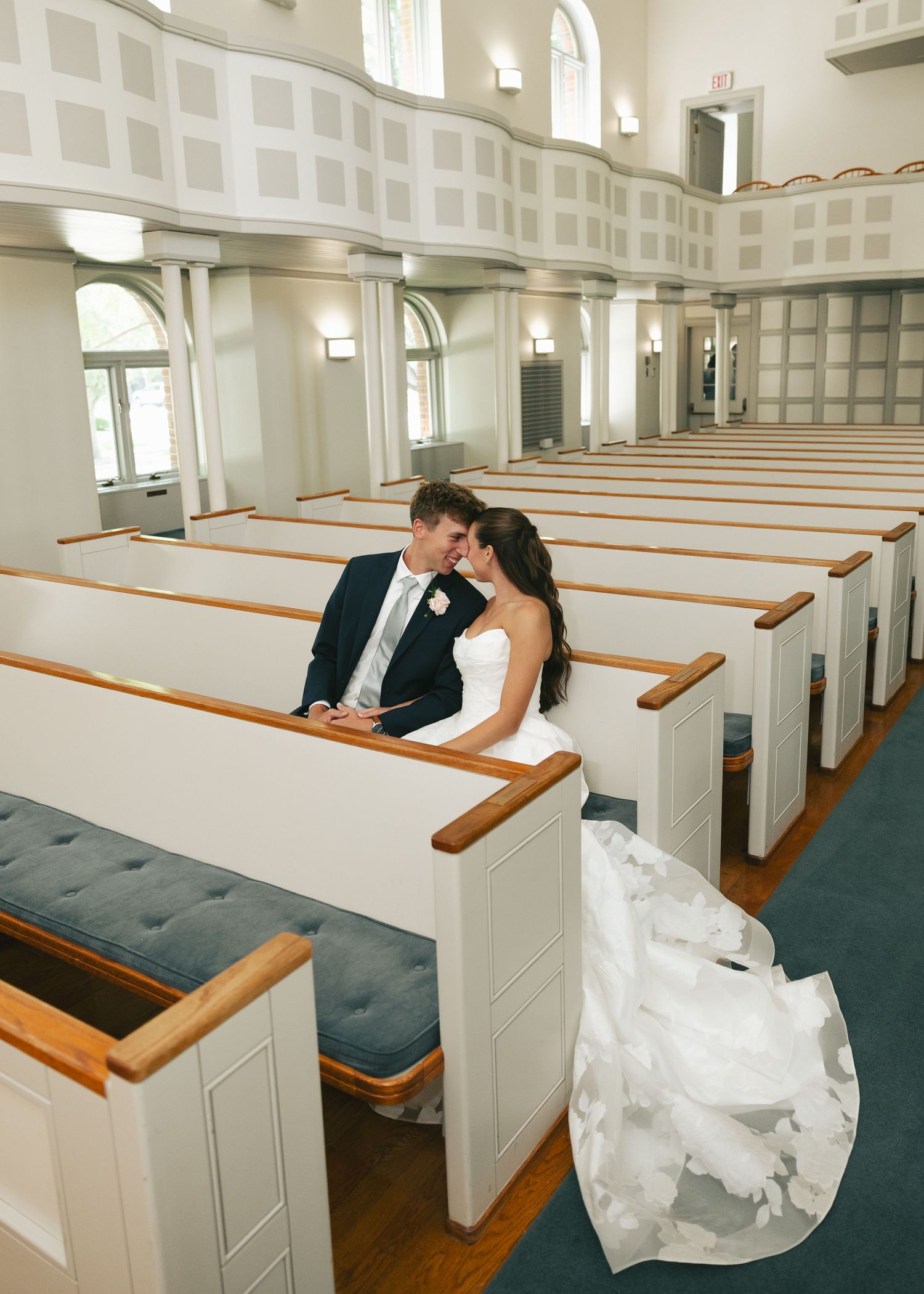 Bride and groom sharing a romantic moment in white church pews, bride in elegant strapless gown with long train.