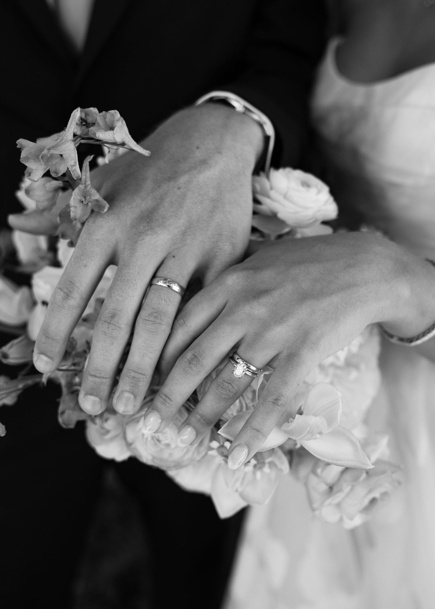 Black and white photo of newlyweds displaying wedding rings on hands resting over a bridal bouquet.