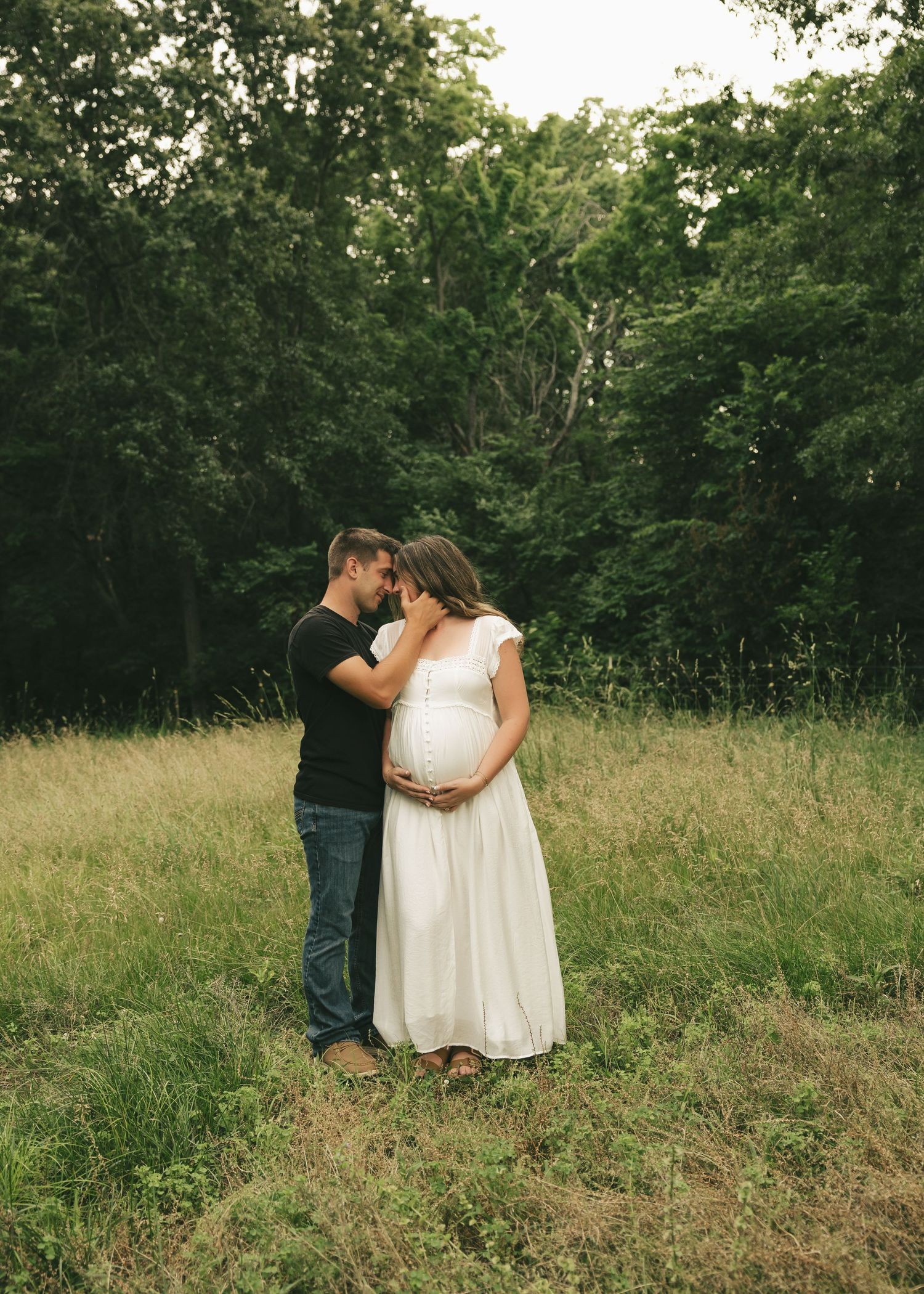 Couple sharing tender moment in grassy field, pregnant woman in white dress with partner in black shirt, lush green trees behind.