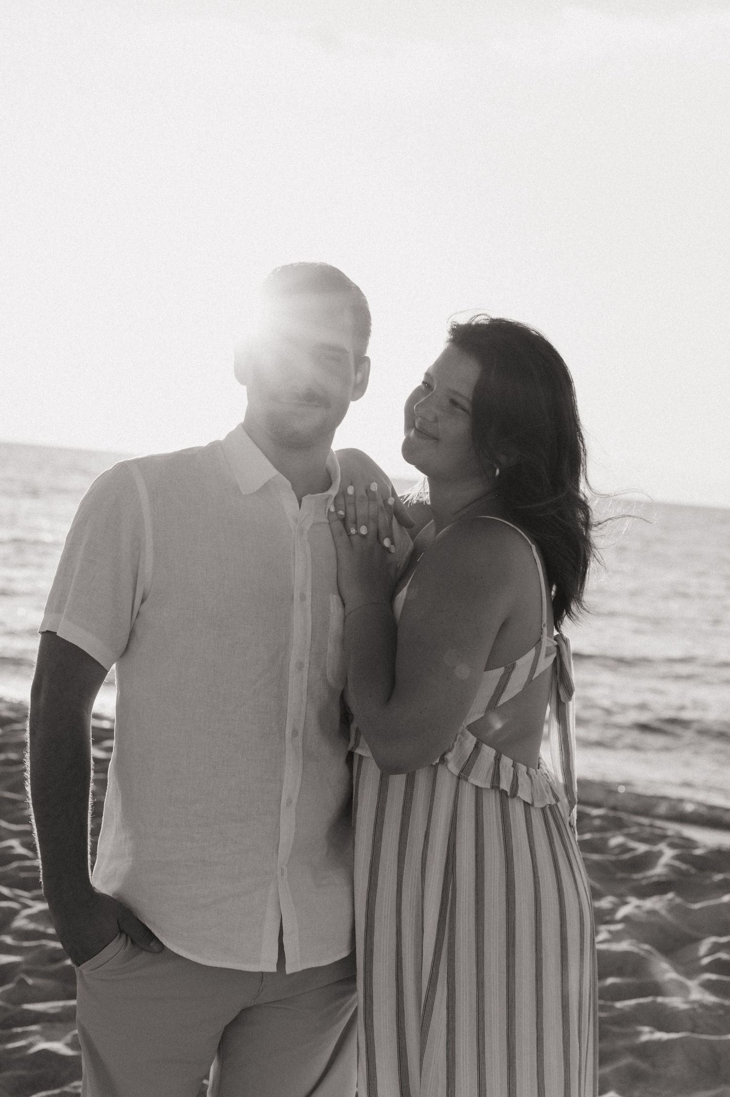 Black and white photo of a couple embracing on a beach at sunset, woman in striped dress laughing joyfully.
