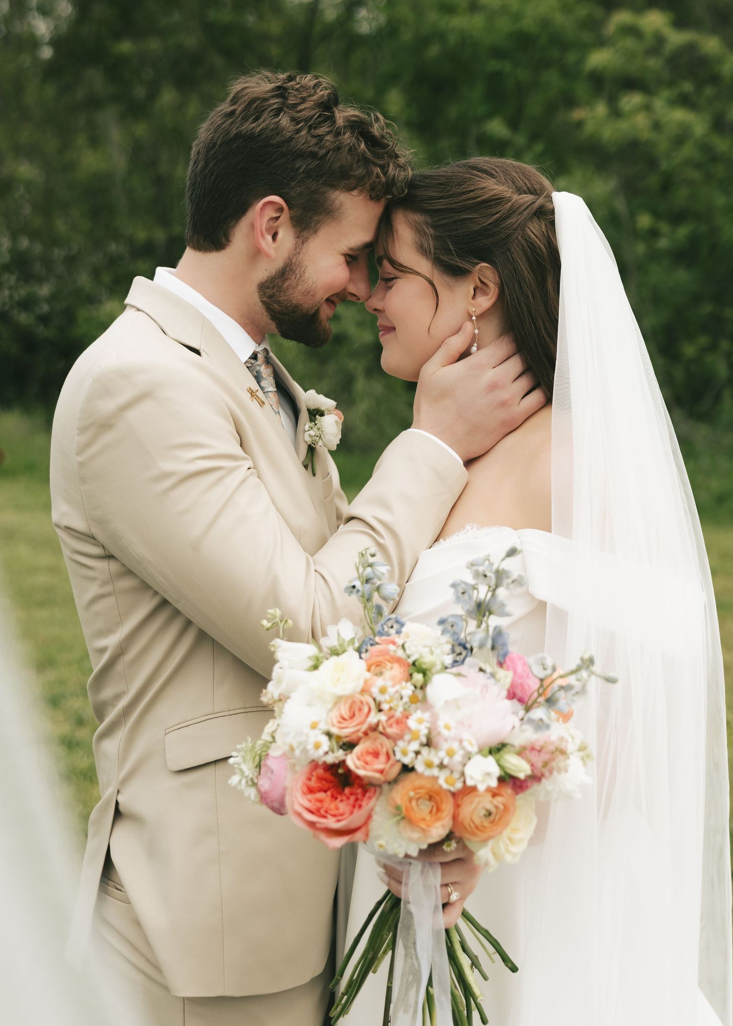 Bride and groom touch foreheads tenderly, bride holding colorful spring bouquet in lush outdoor setting.