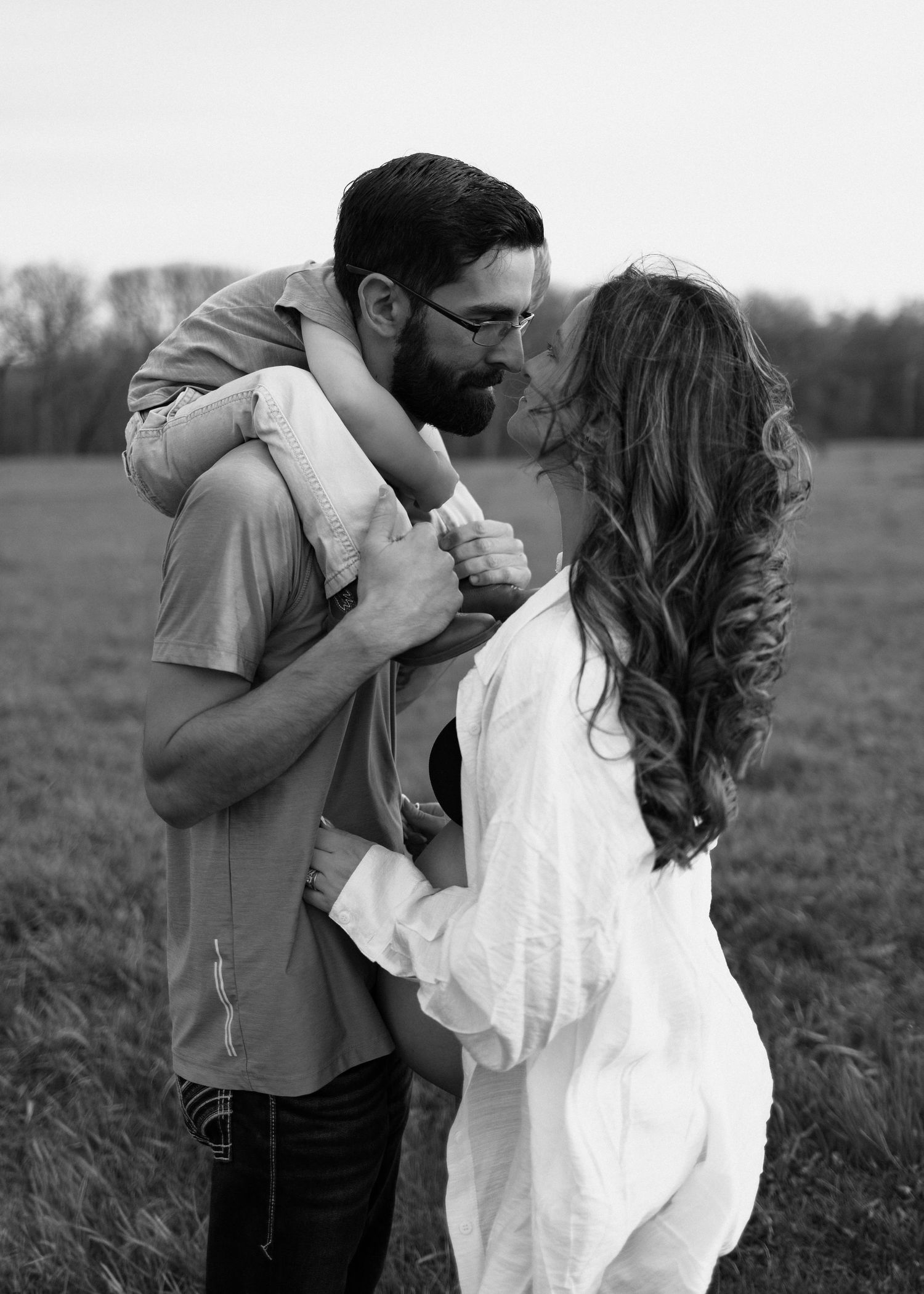 Black and white photo of a couple sharing a tender moment in a field while holding their baby.