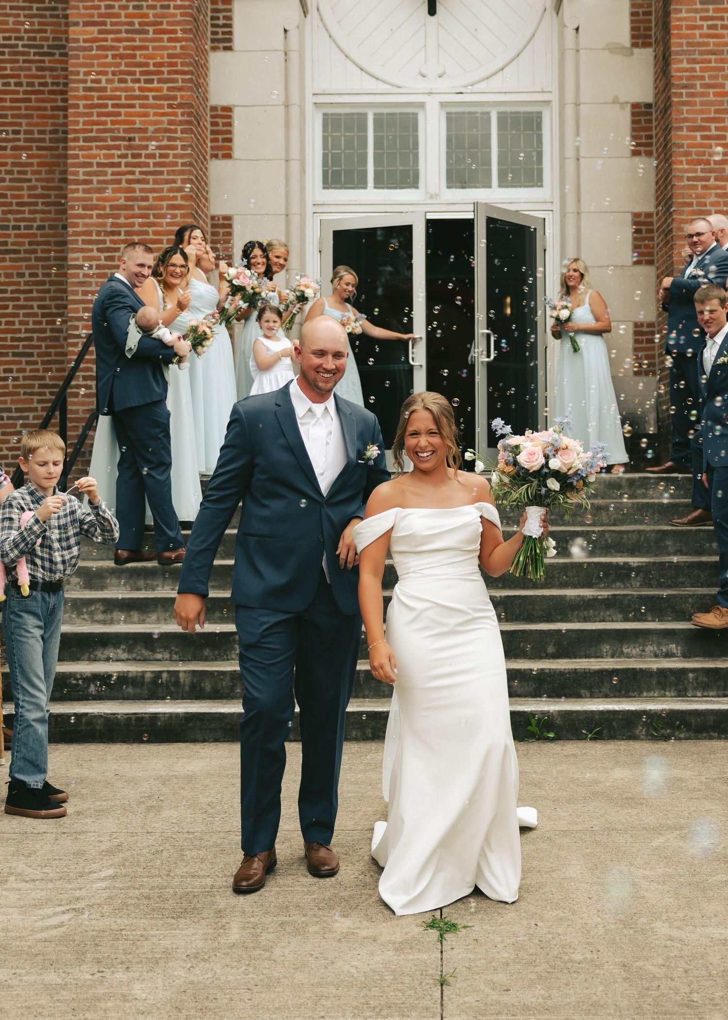 Bride and groom exit brick church amid confetti, surrounded by wedding party in navy suits and blue bridesmaid dresses.