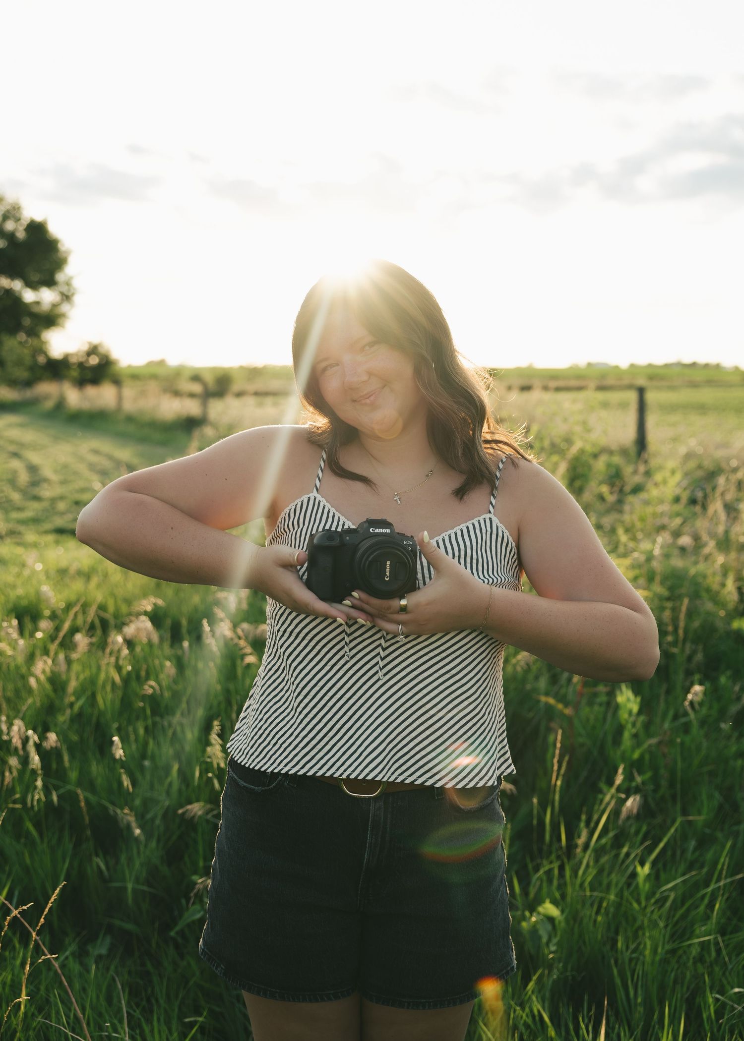 Woman holding a DSLR camera smiling in a sunlit green field at golden hour.