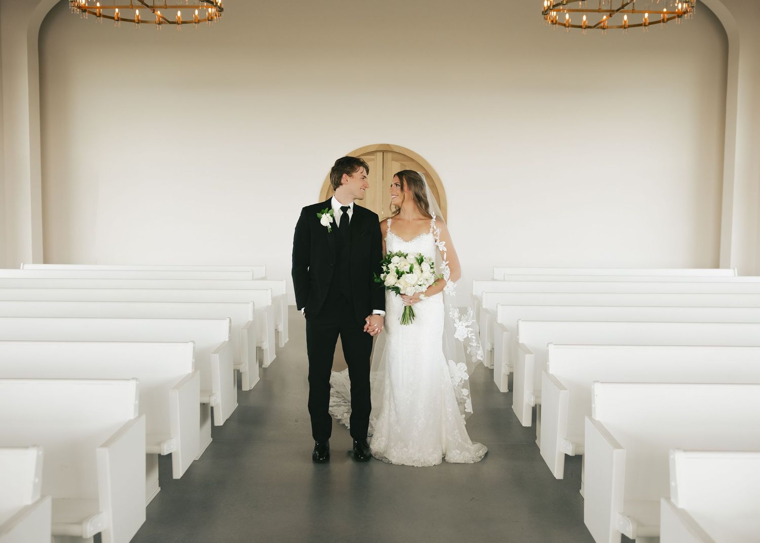 Bride and groom stand hand in hand at the altar of a modern white chapel with rows of white pews.
